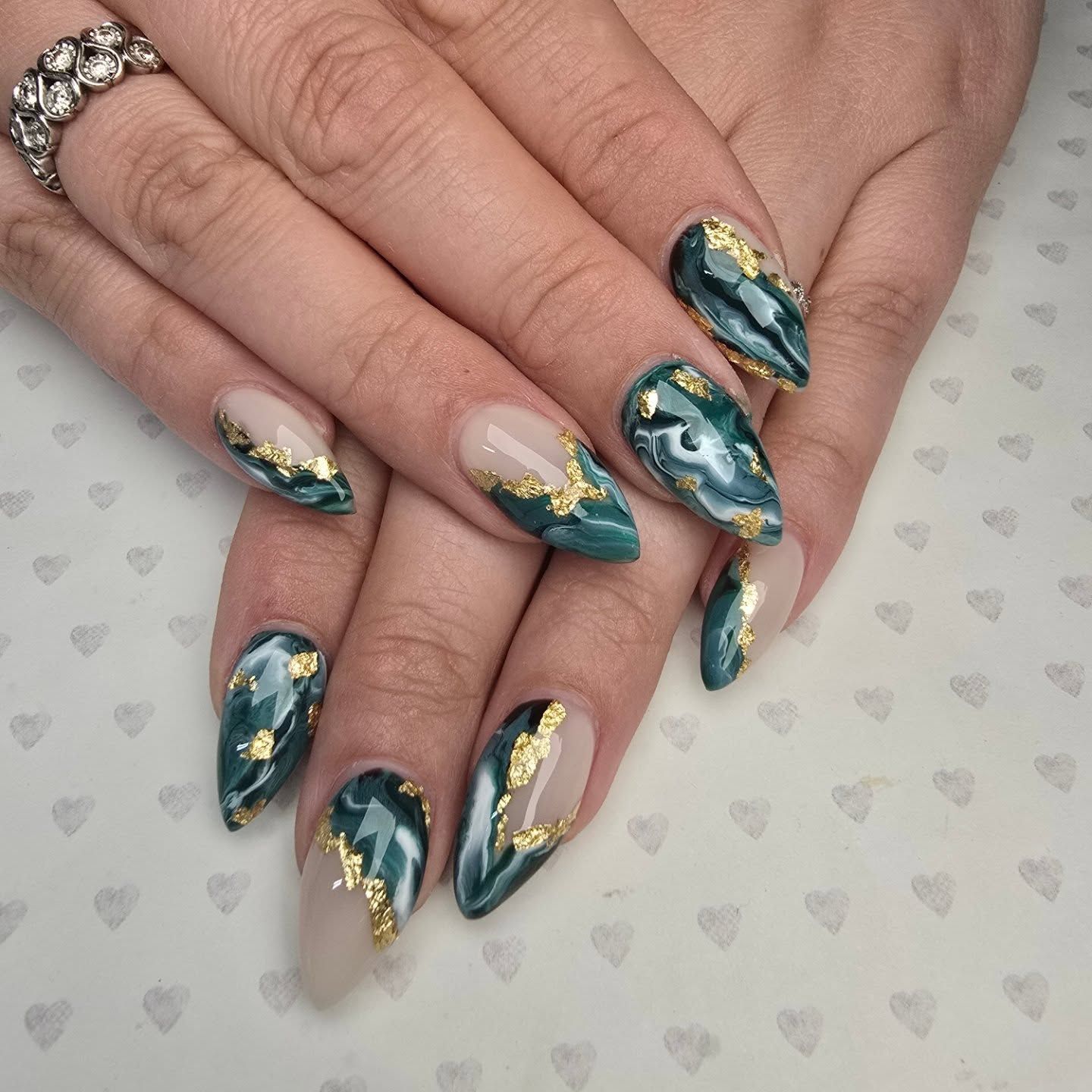 Stiletto nails with teal marble design and gold foil accents on a white heart-patterned background offered for bride.