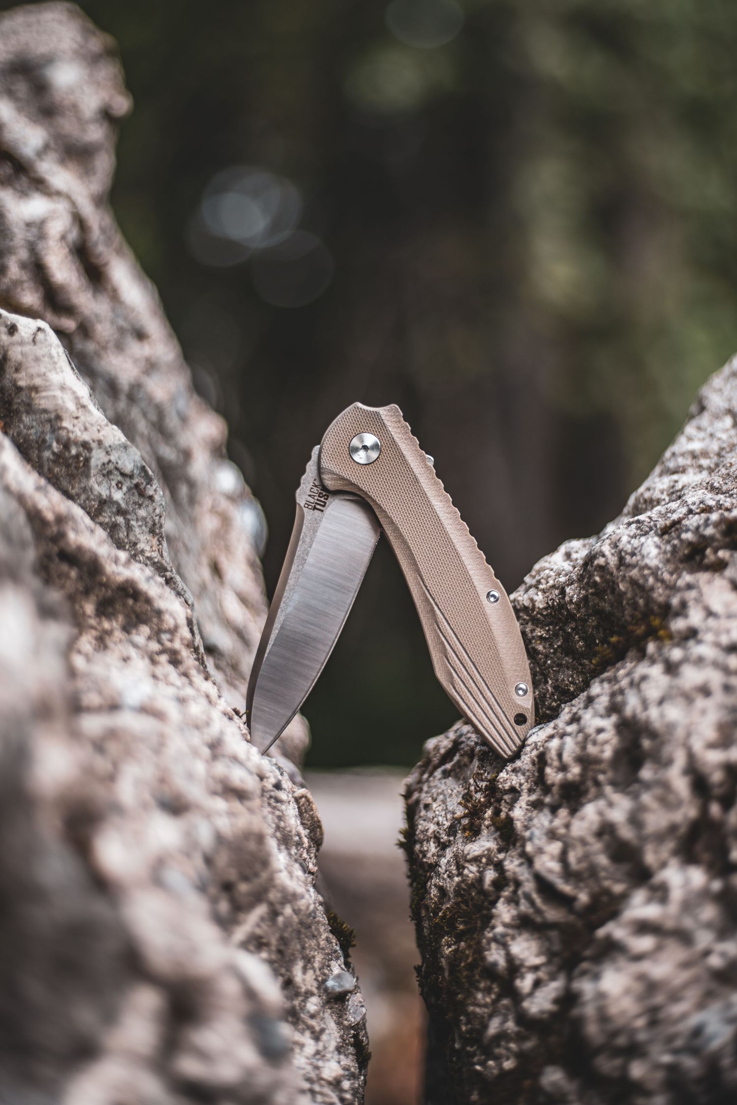 A folding pocket knife with a tan handle balanced open between two rocks outdoors. Product Photography in Prince George, BC