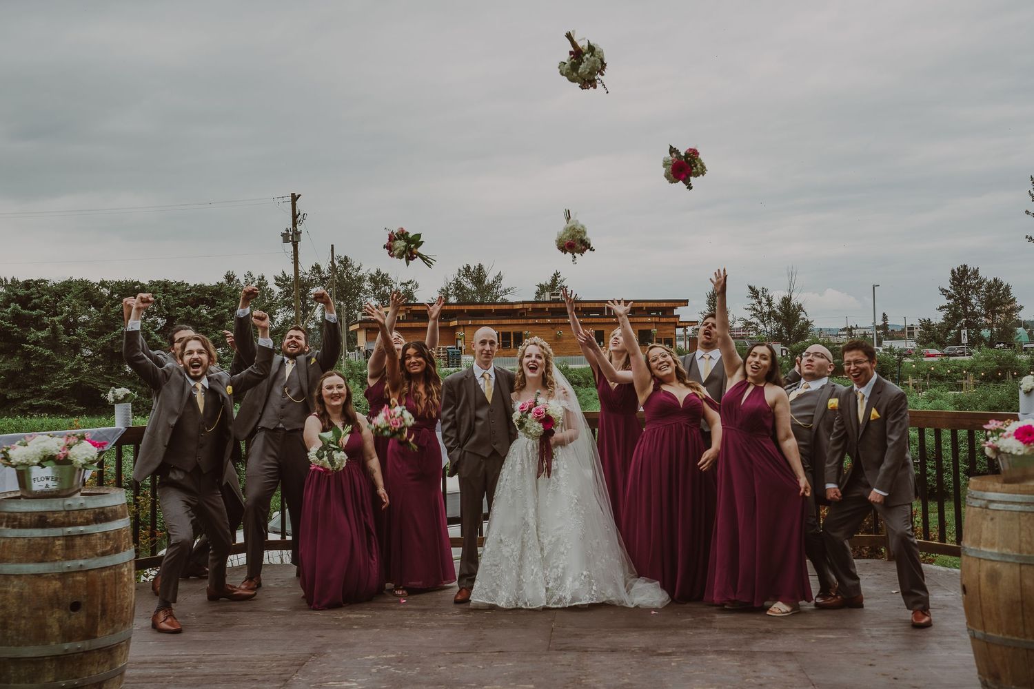 Wedding party in burgundy and gray celebrates by tossing bouquets on a rustic outdoor venue deck at Northern Lights Winery, Prince George.