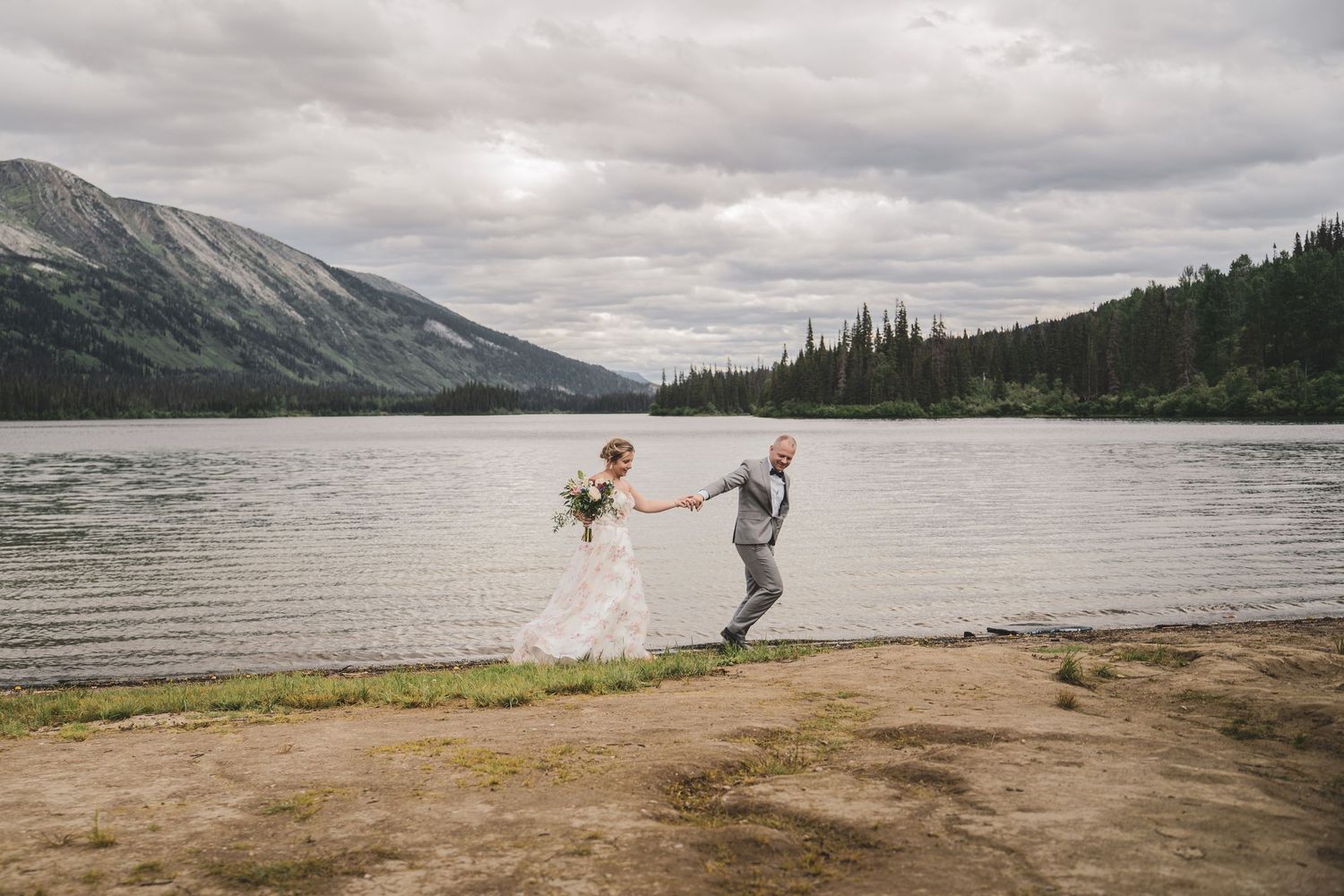 Bride and groom hold hands and run playfully along the shore of a mountain lake on their wedding day.