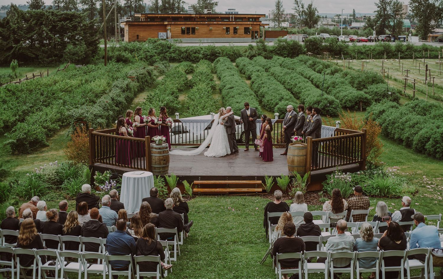 Outdoor wedding ceremony at a vineyard with guests seated on white chairs facing a wooden deck altar.