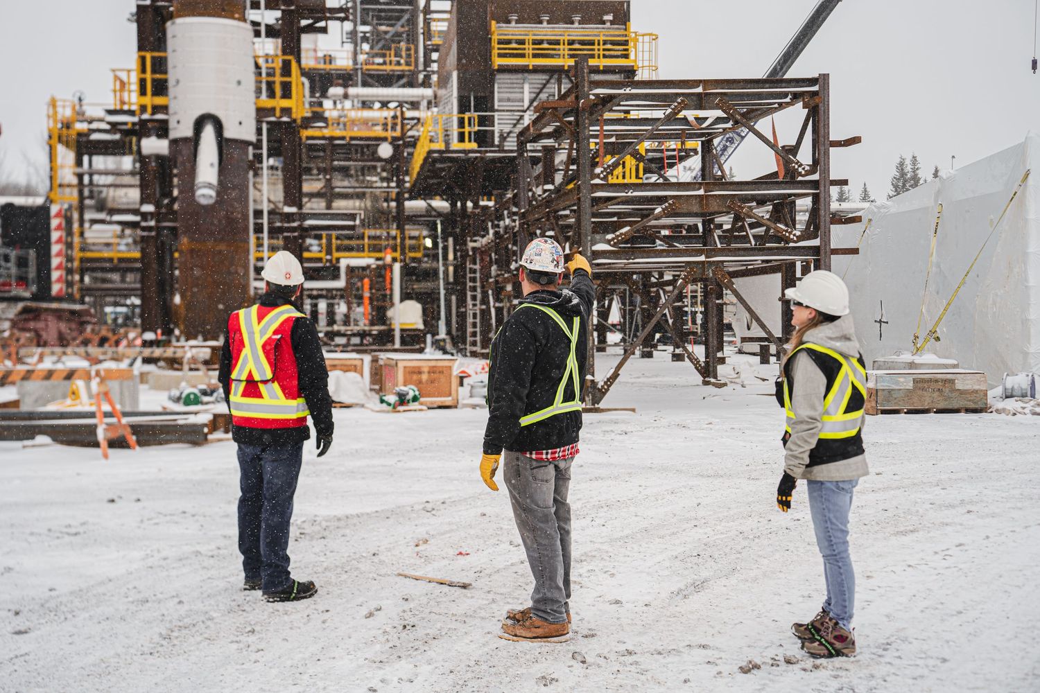 Three construction workers in safety vests and hard hats standing in a snowy industrial site.