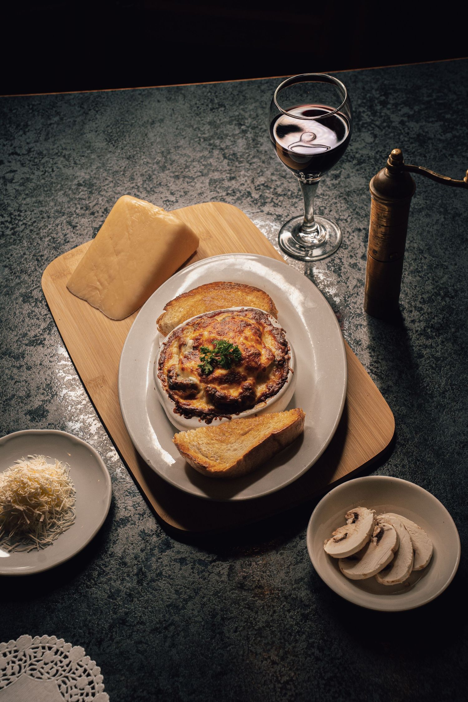 Baked moussaka on a wooden board with toast, red wine, sliced mushrooms, and grated Parmesan cheese.