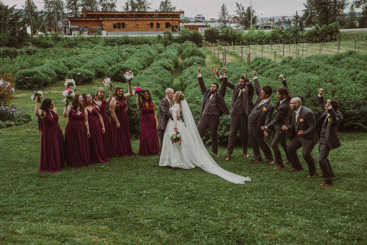 Wedding party celebrates joyfully in a lush green field with bridesmaids in burgundy dresses at Northern Lights Winery venue in PG.