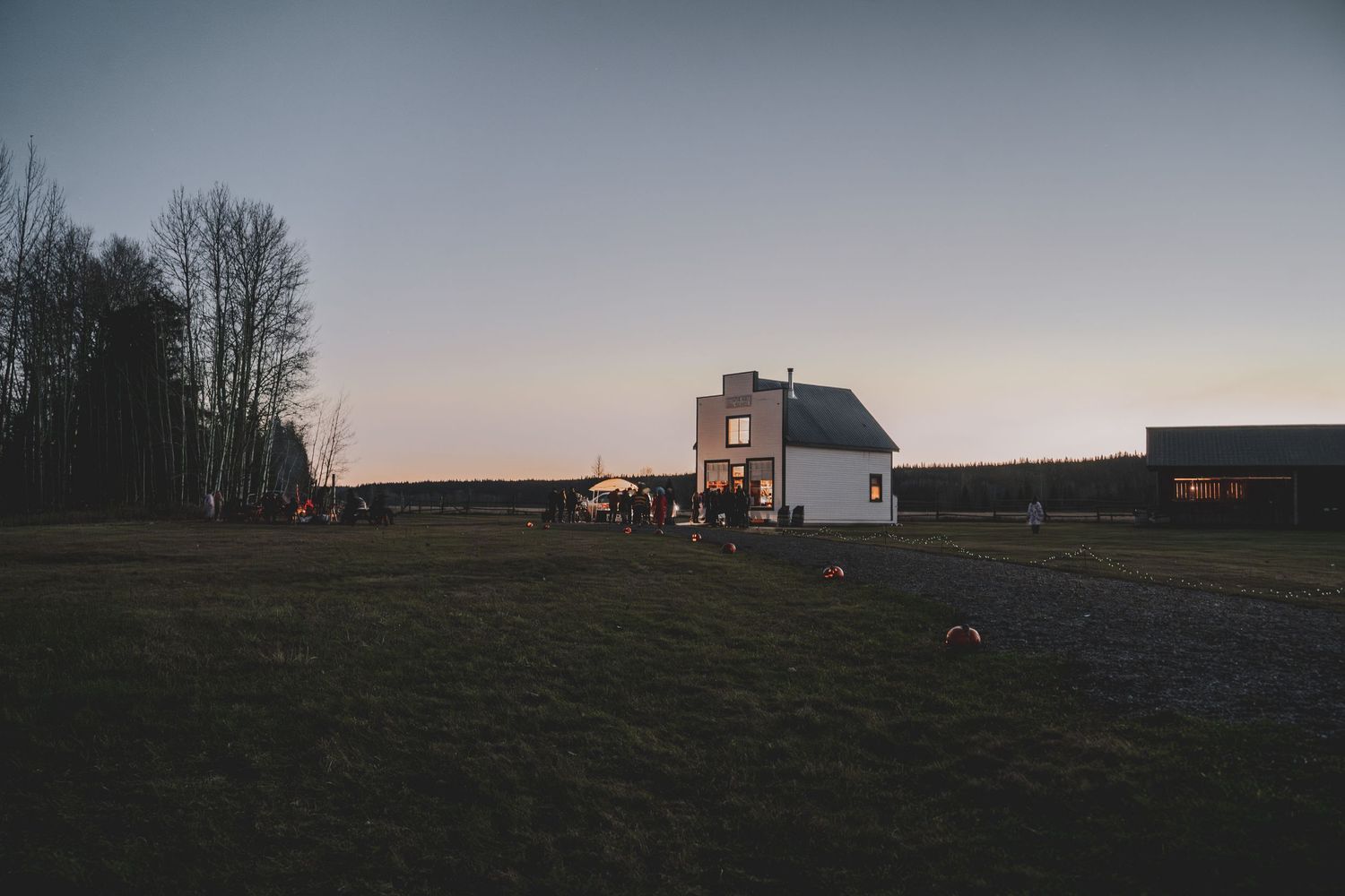 White farmhouse glows at dusk surrounded by open fields with guests gathered for an evening event at Huble Homestead in Prince George, BC.