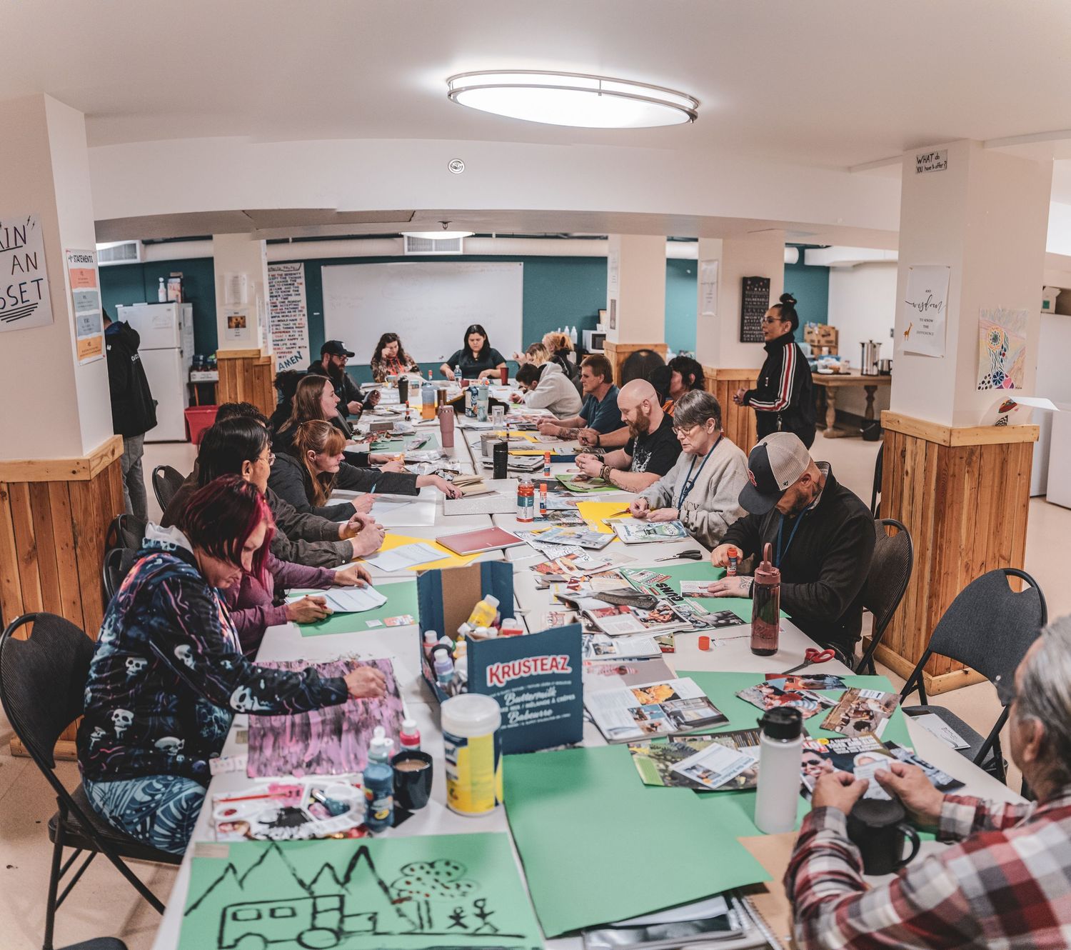 Large diverse group of people participate in an art workshop around long tables in a community center.