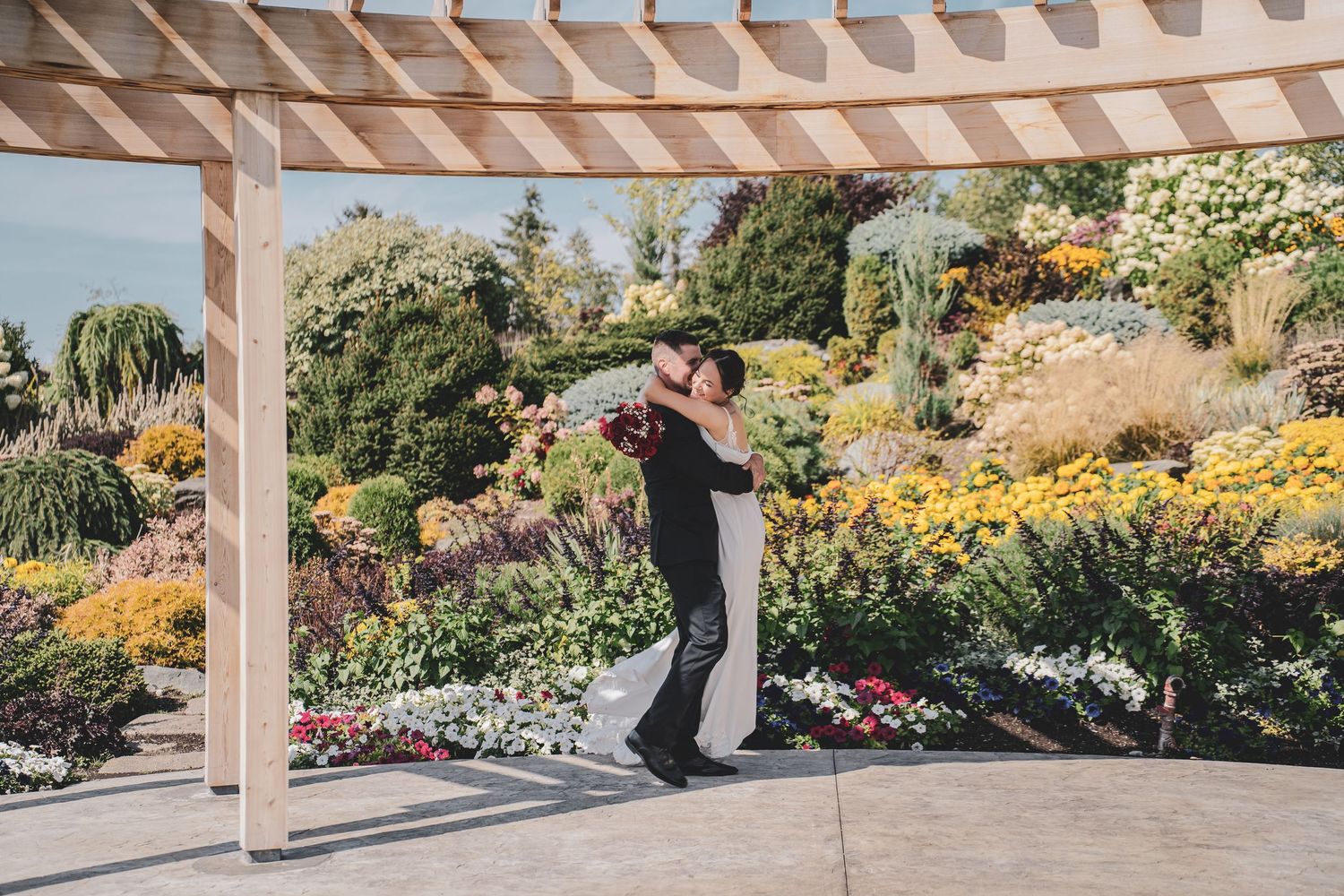 Bride and groom dancing under pergola surrounded by vibrant colorful garden in bloom.