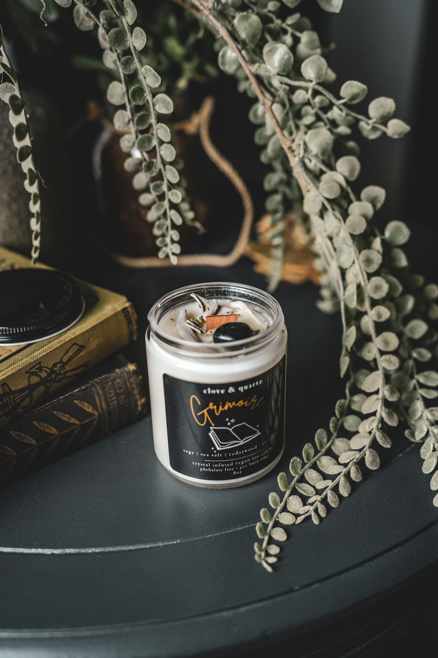 Product photography of Grimoire scented soy candle in glass jar with botanicals on dark table with eucalyptus.