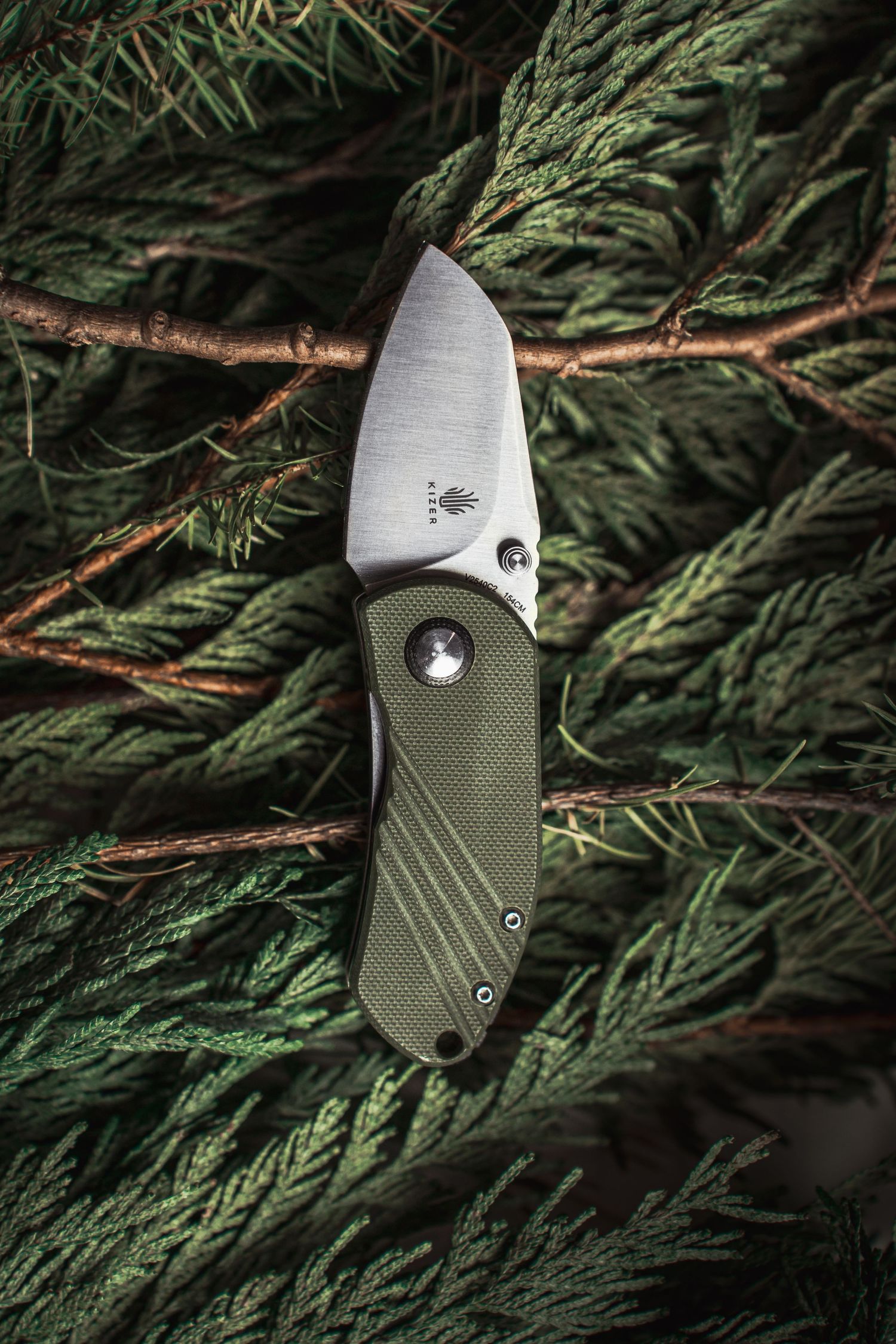 Product Photography. of a green handled folding pocket knife with brushed steel blade resting on evergreen pine branches.