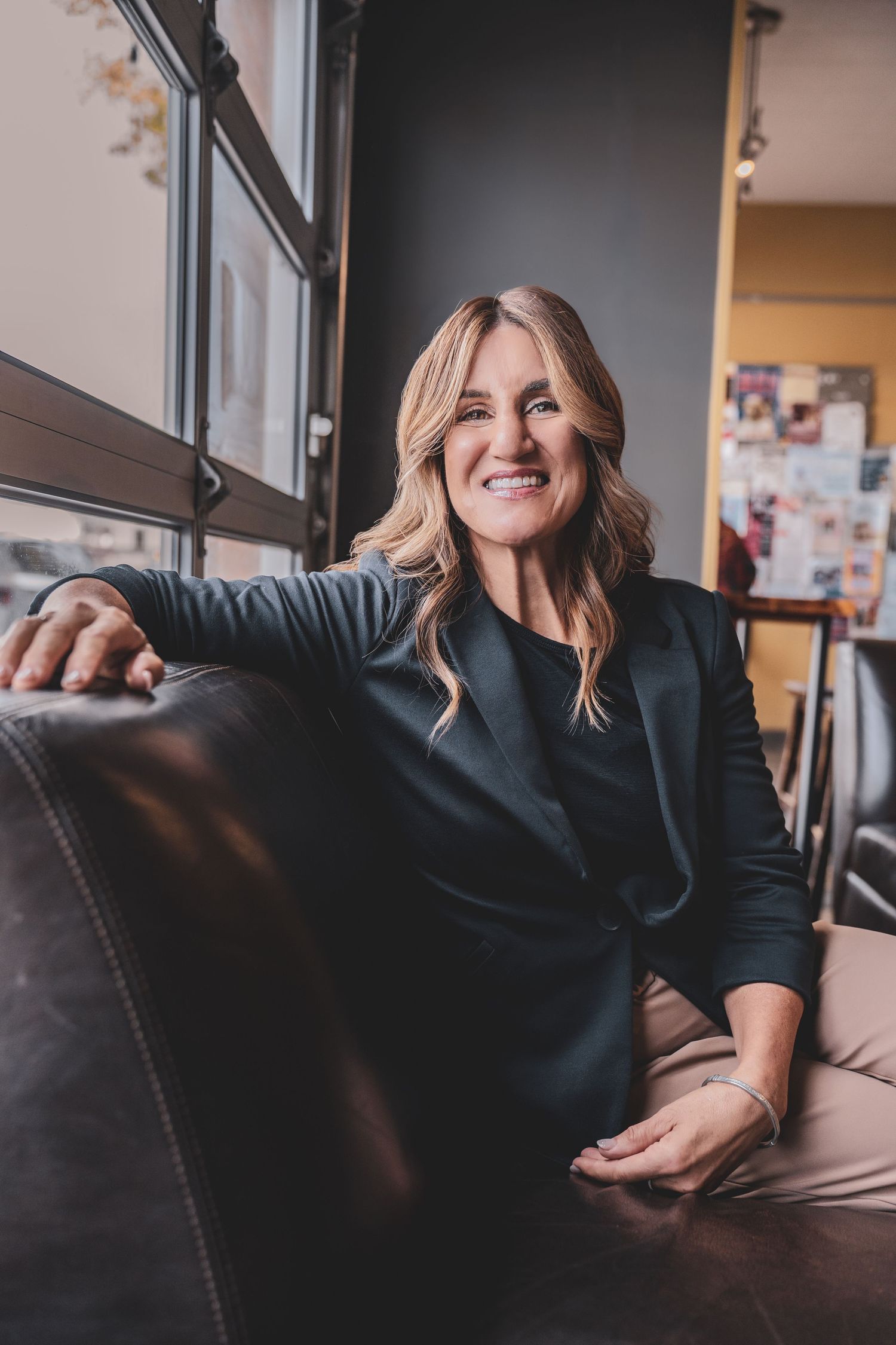 Confident blonde businesswoman smiling while relaxing on a leather sofa near a window.