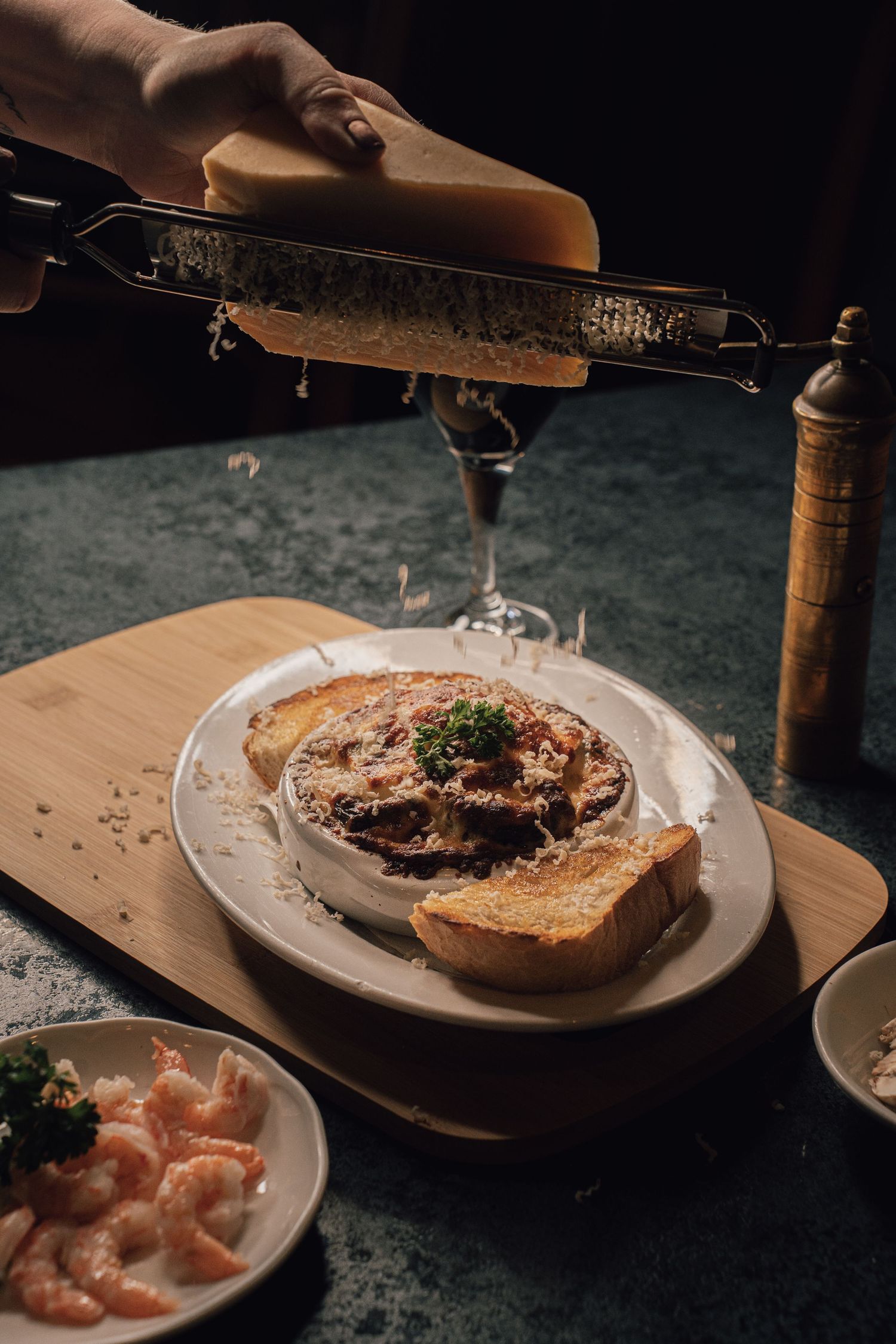 Hand grating Parmesan cheese over a baked moussaka dish on a wooden board with shrimp on the side.