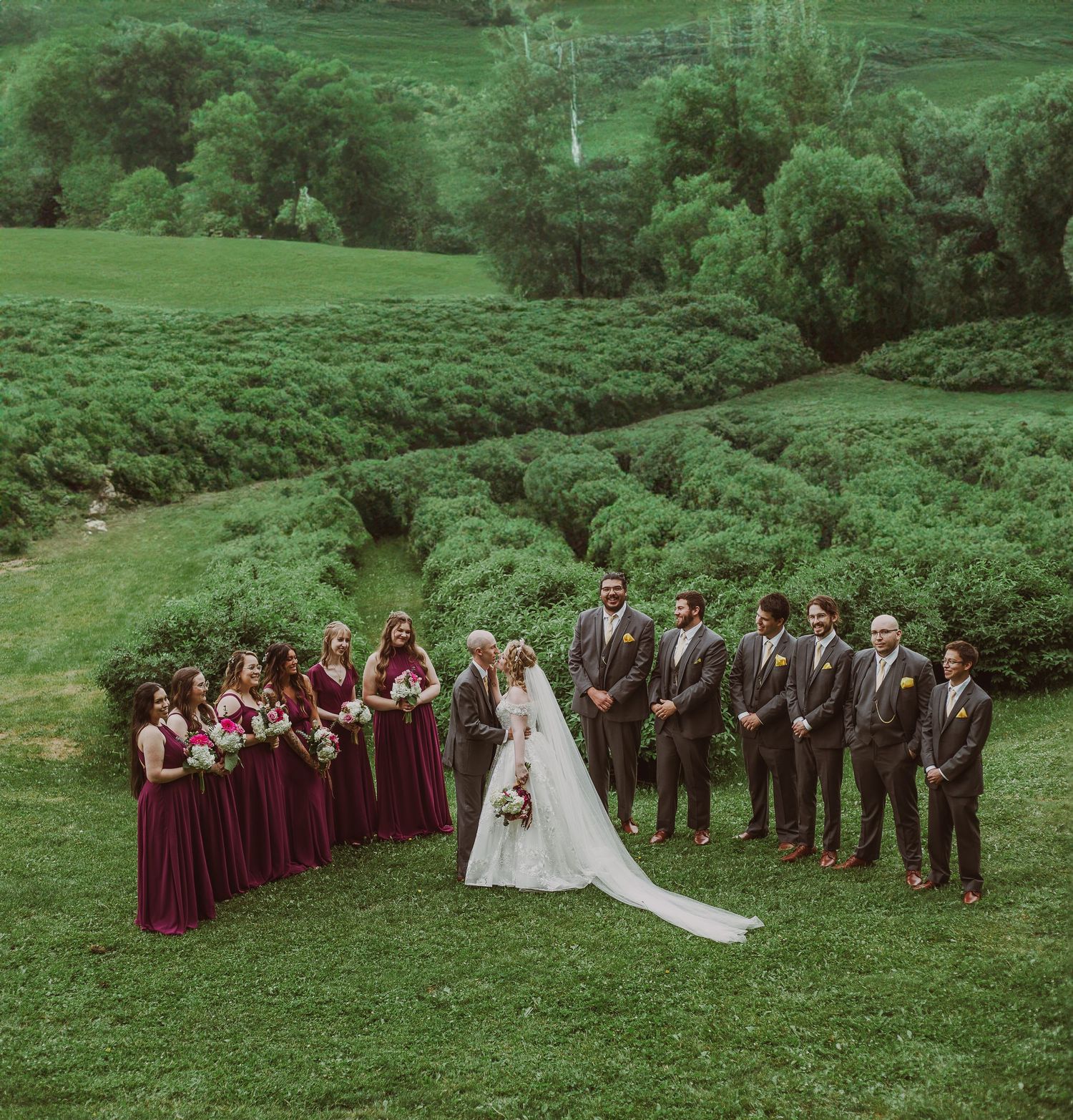 Aerial view of wedding party in burgundy and grey standing beside lush green terraced garden rows at Northern Lights Winery, Prince George,