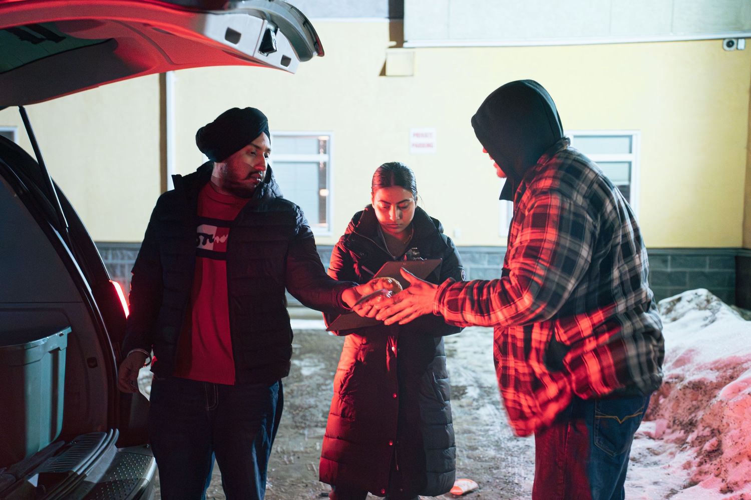 Three people in winter clothing gather around an open car trunk in a snowy parking lot during outreach work for Prince george, bc.