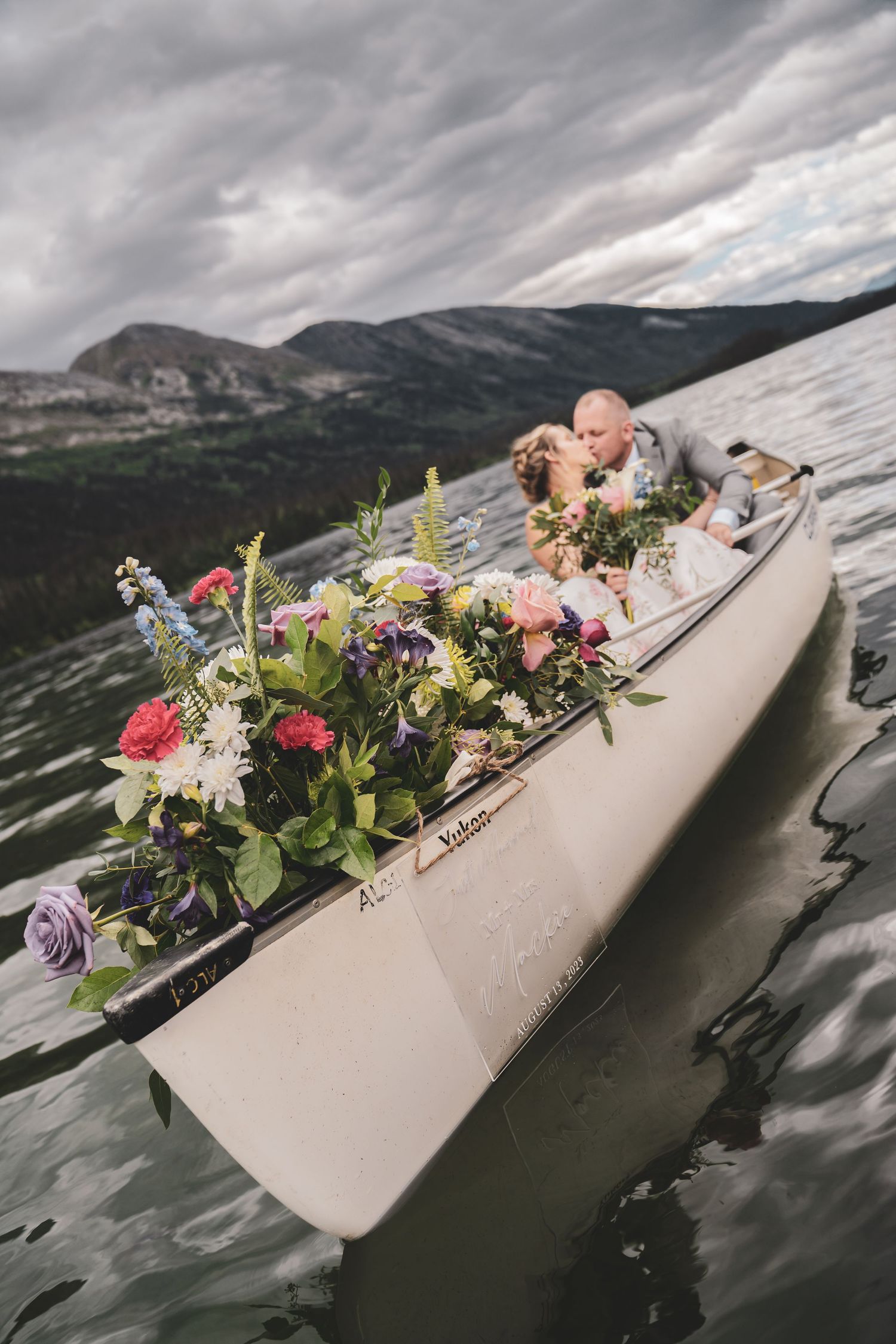 Bride and groom kissing in a flower-filled canoe on a scenic mountain lake under cloudy skies in the Pine Pass, BC offered by Free West.