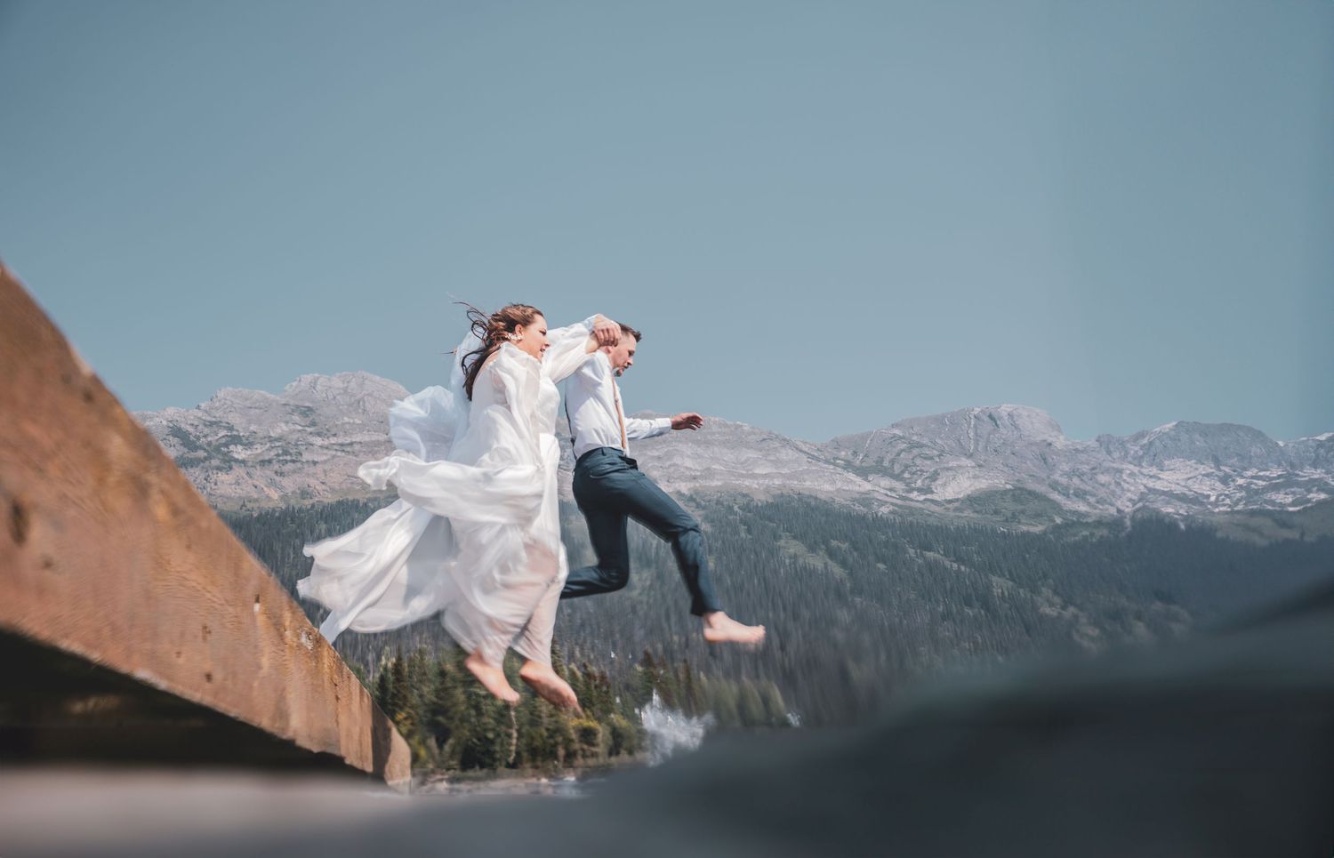 Bride and groom joyfully jumping off a wooden dock with stunning mountain scenery behind them as they complete their vows at elopement.