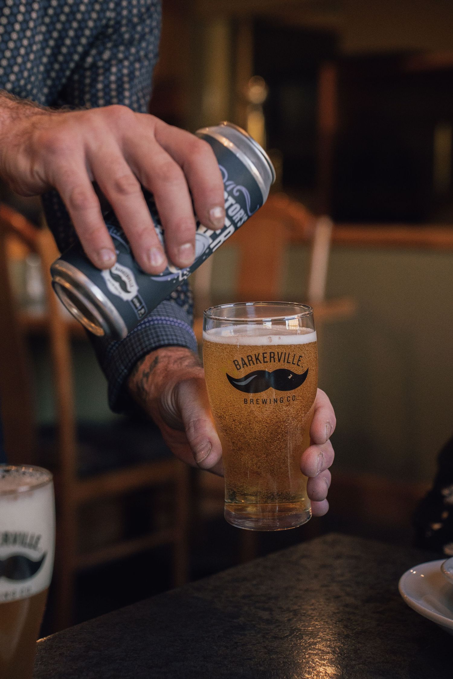 Person pouring Barkerville Brewing craft beer from a can into a branded pint glass at a bar.