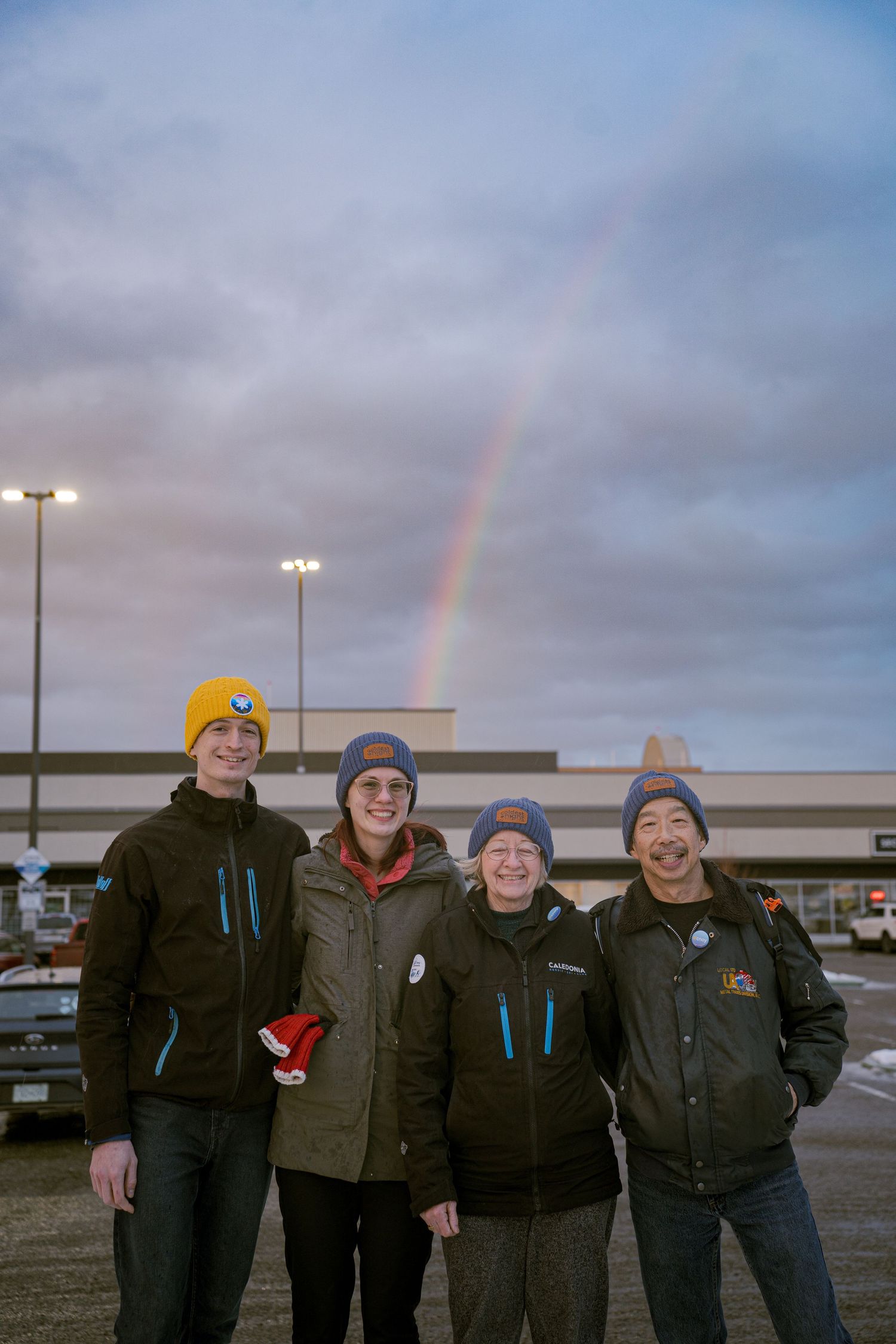 Four people in winter hats smile outdoors with a rainbow visible in the cloudy sky behind them.