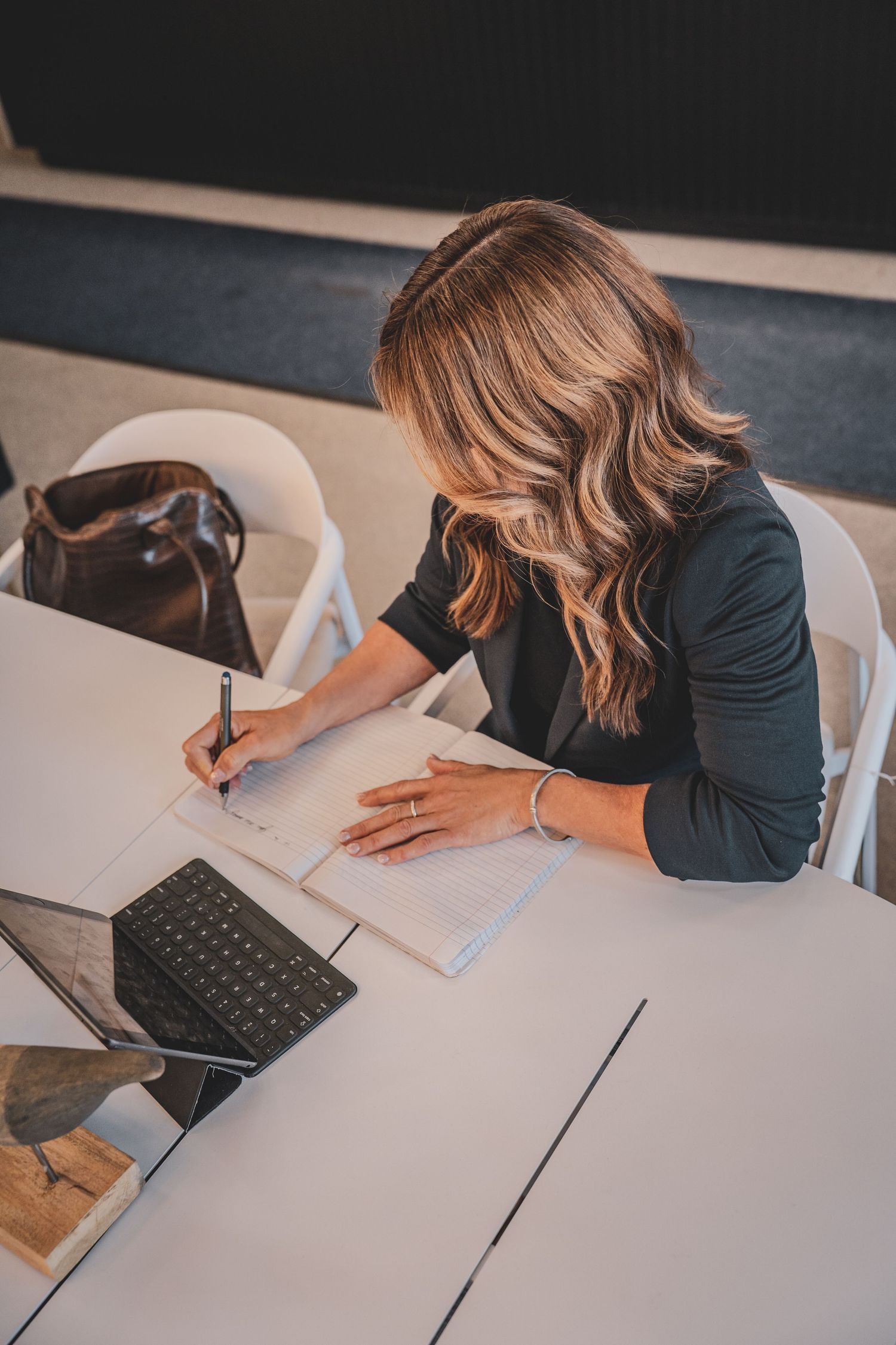 Businesswoman writing notes in a notebook beside a tablet at a white conference table.