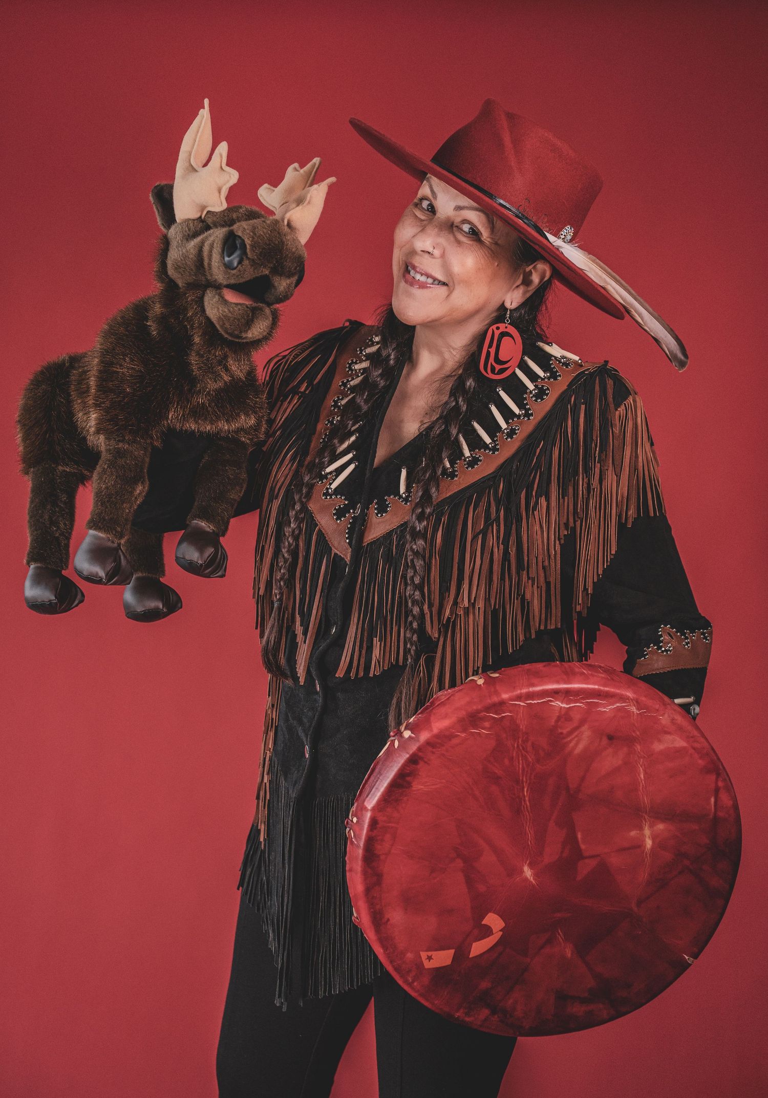 Kym Gouc red hat holding moose puppet and drum in colorful Indigenous-inspired performance outfit during musician portrait photoshoot.