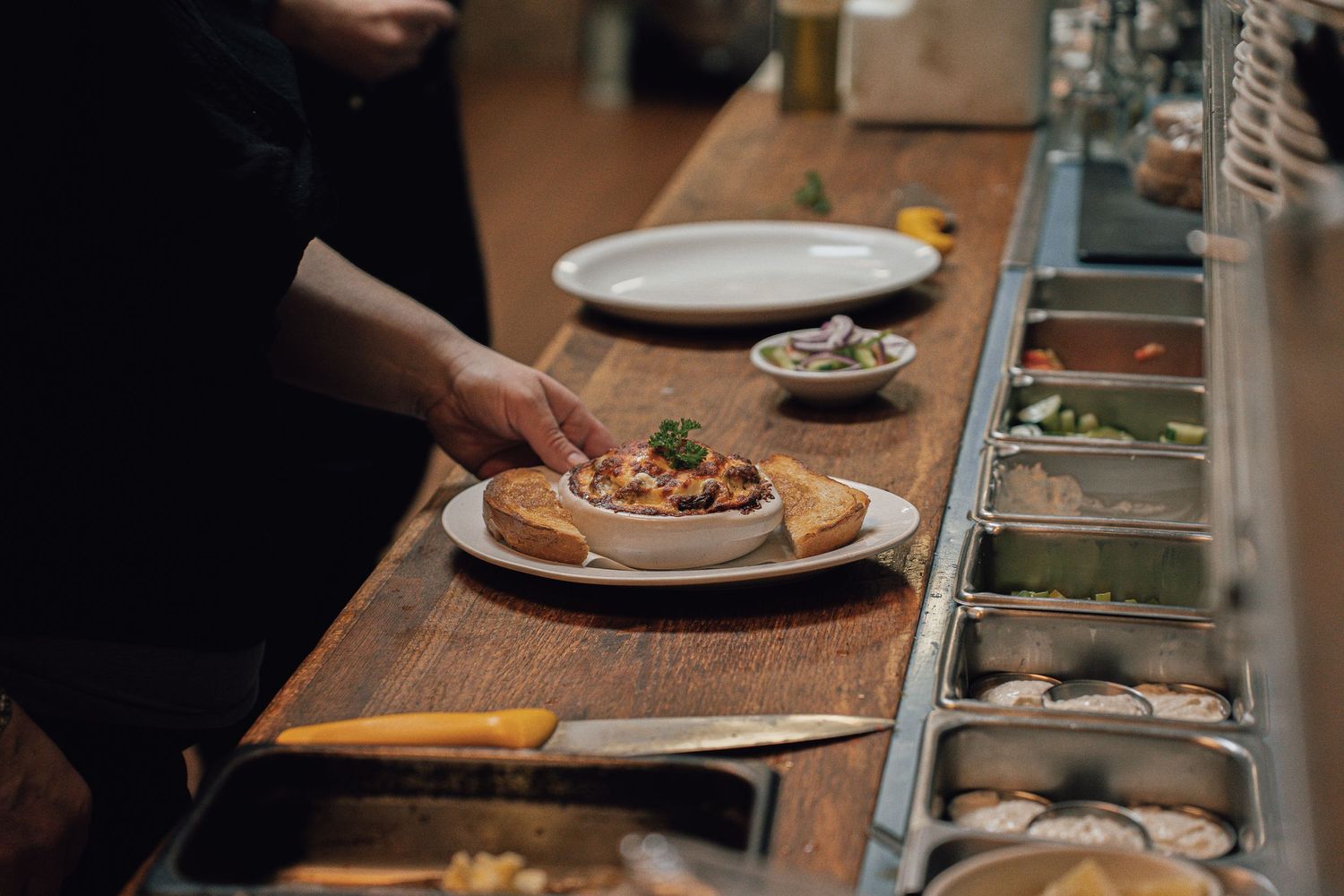 Chef plating a gourmet open-faced sandwich with toppings on a wooden prep counter in a professional kitchen.