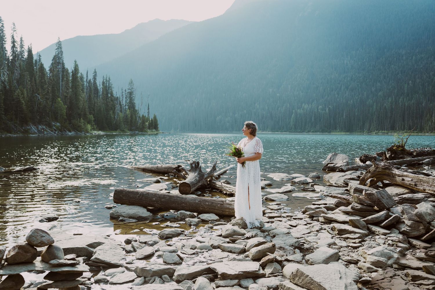 Bride holding bouquet standing alone on rocky mountain lake shore with forested peaks behind her.