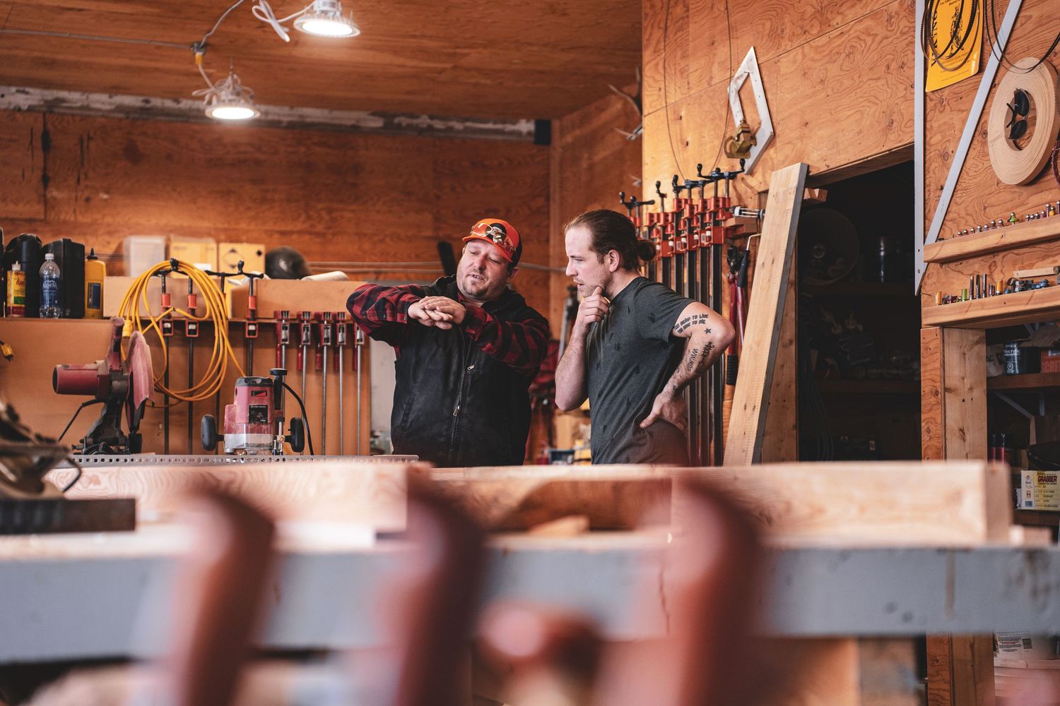 Two craftsmen discussing woodworking projects in a rustic workshop filled with tools and lumber.