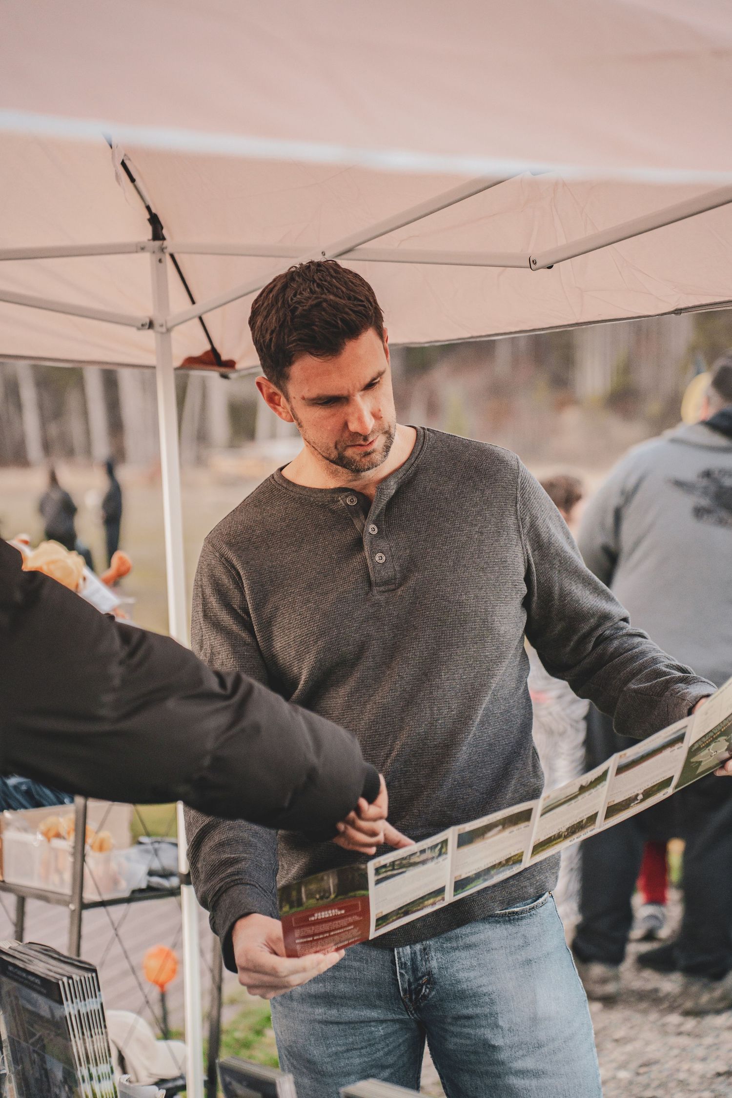 Man in gray sweater browses printed materials at an outdoor vendor booth under a white canopy tent for tourism prince george event.
