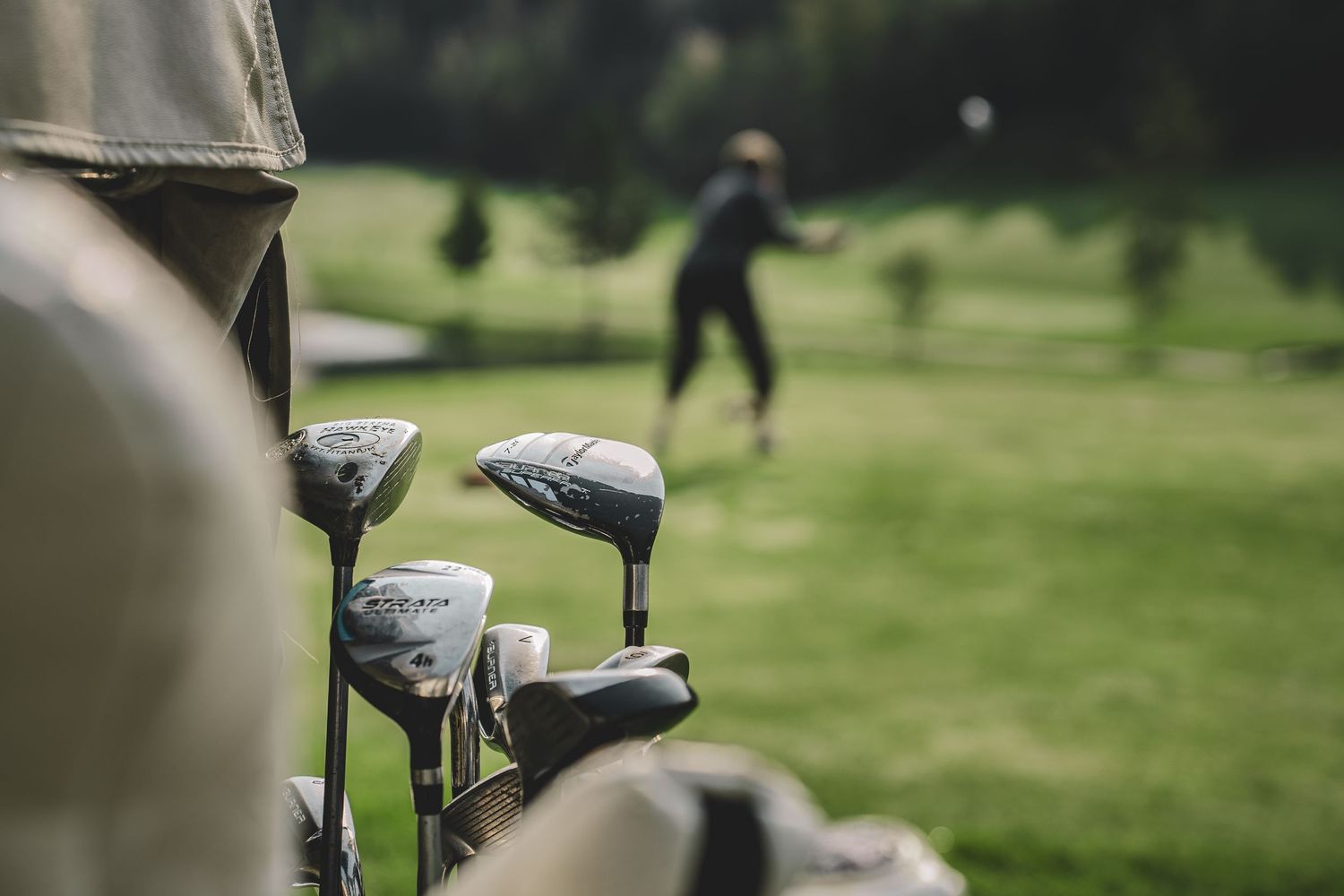 Golf clubs in a bag foreground with a golfer swinging on a lush green golf course in the background.