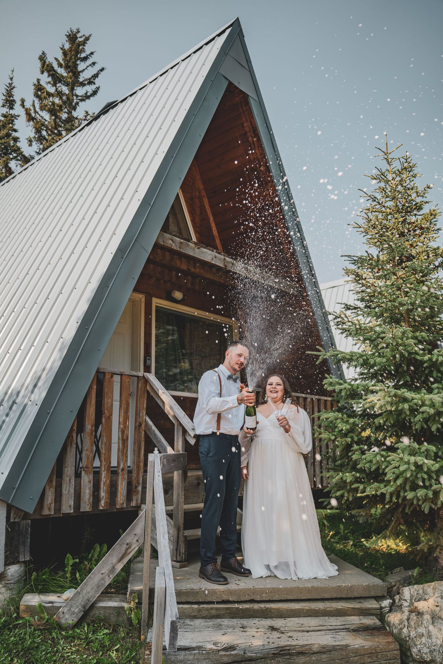 Bride and groom celebrate with champagne spray outside an A-frame cabin surrounded by evergreen trees.