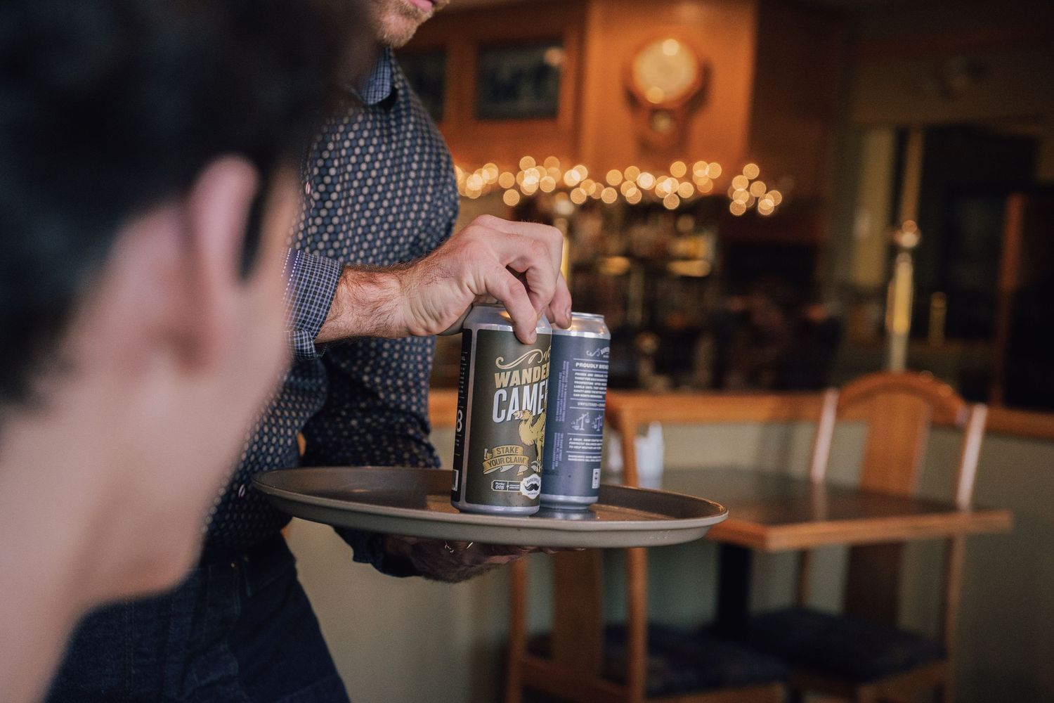 Waiter serving canned drinks on a round tray in a warmly lit restaurant.