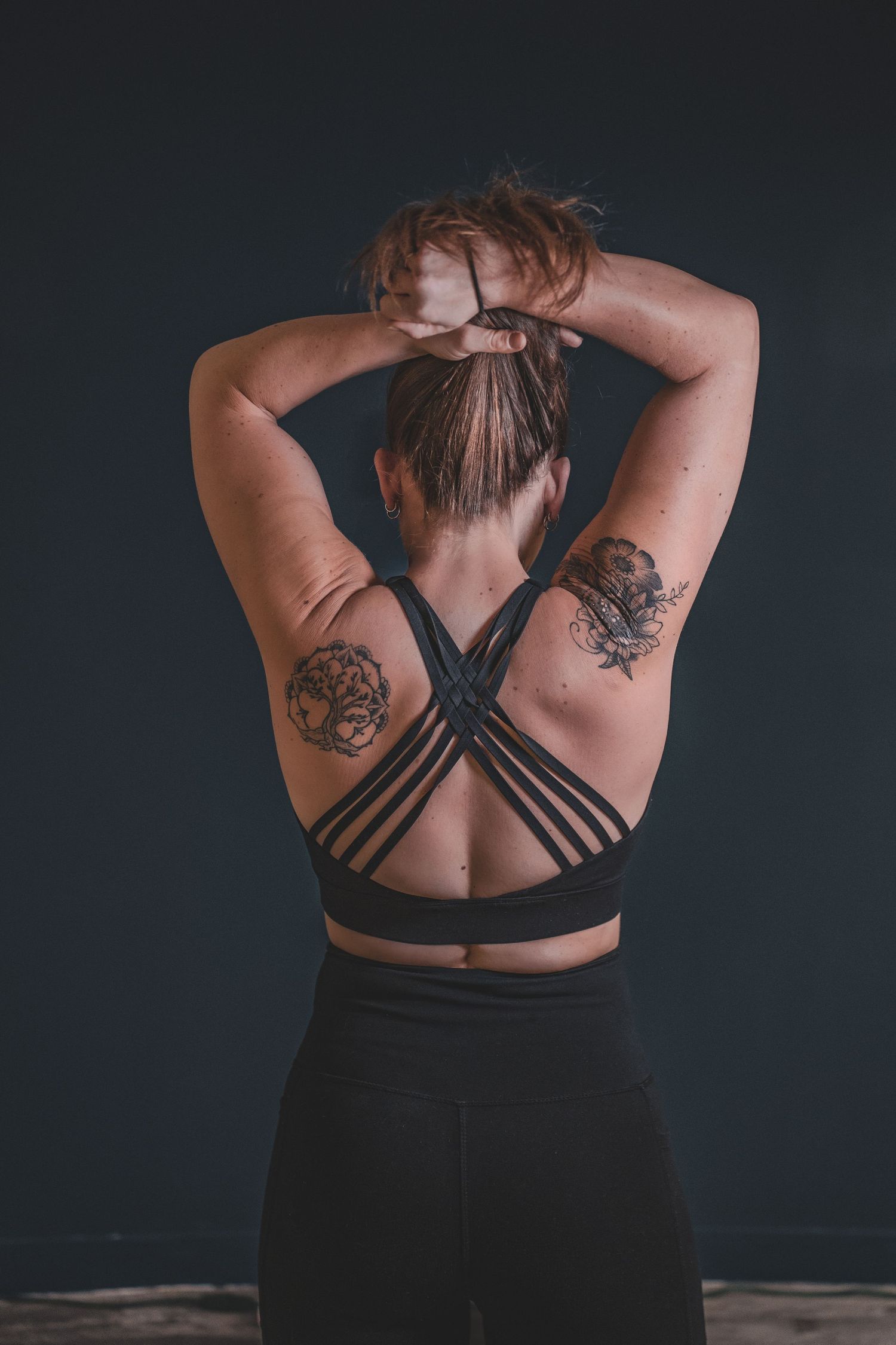 A fit woman with tattoos on her back poses in a black strappy sports bra with hands behind her head during a branding photoshoot.