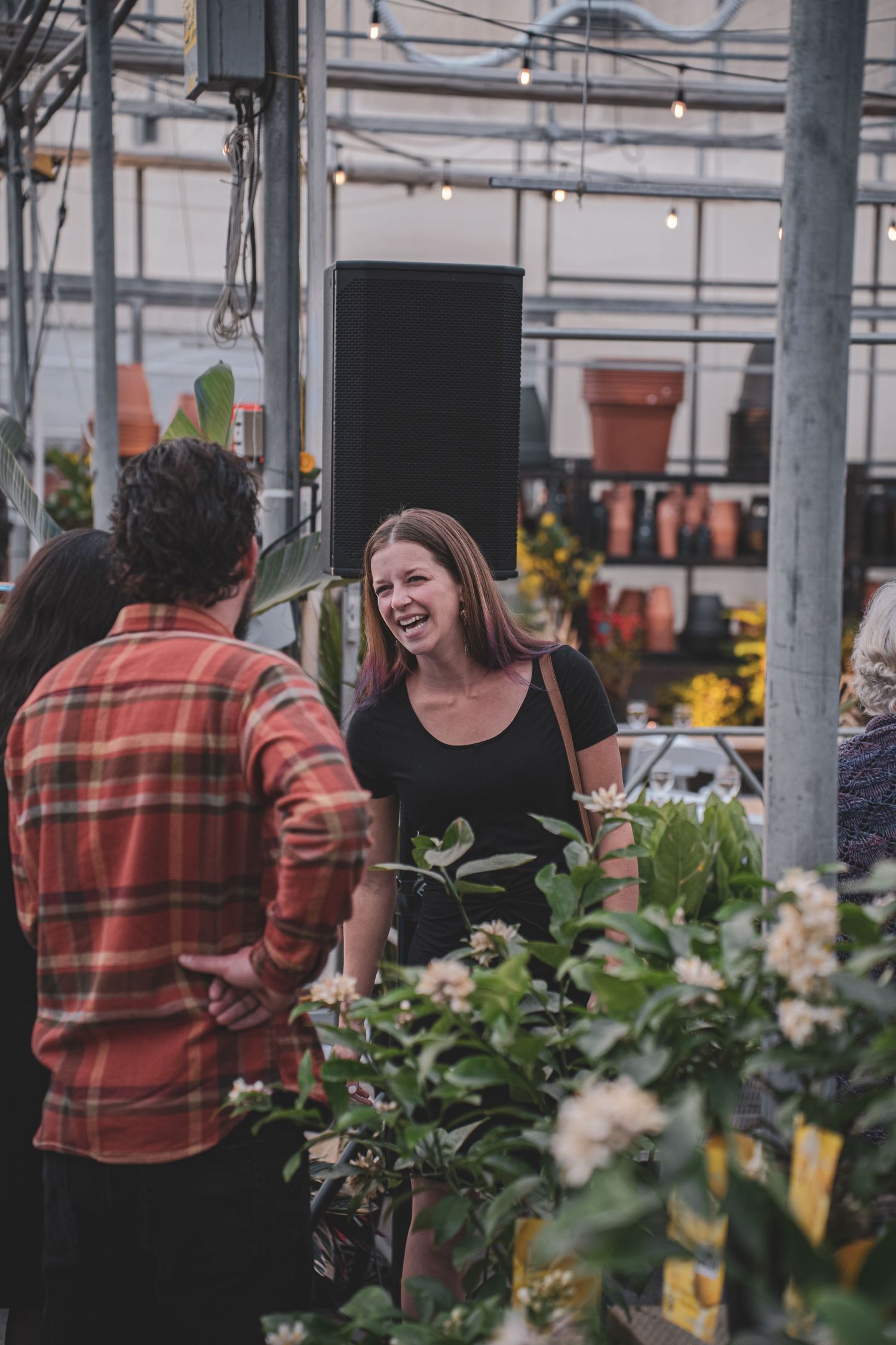 A laughing woman in a black top chats with a man in a greenhouse plant shop surrounded by lush greenery.
