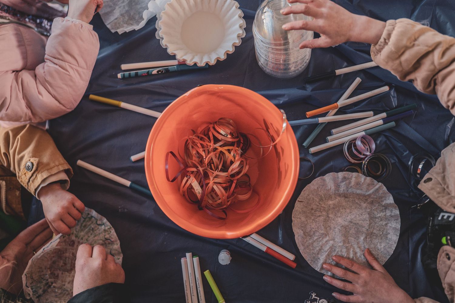 Children engage in a hands-on craft activity with an orange bucket, pencils, and art supplies on a dark table.