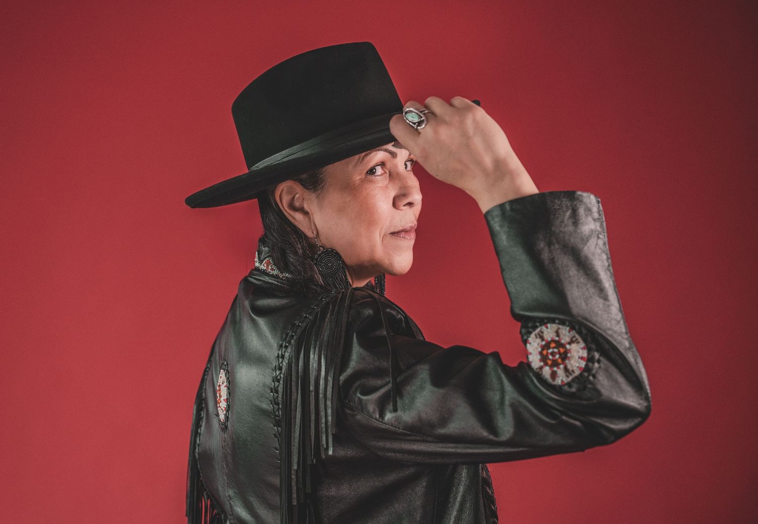 Kym Gouchie in black leather jacket and black cowboy hat posing against vibrant red background during artist photoshoot.