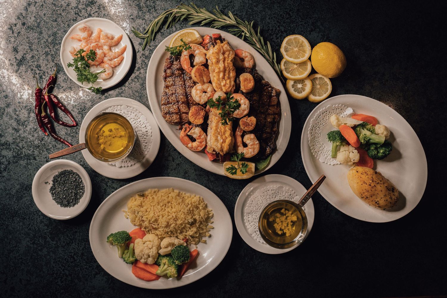 Overhead view of a full seafood and steak dinner spread with sides, sauces, and fresh ingredients.