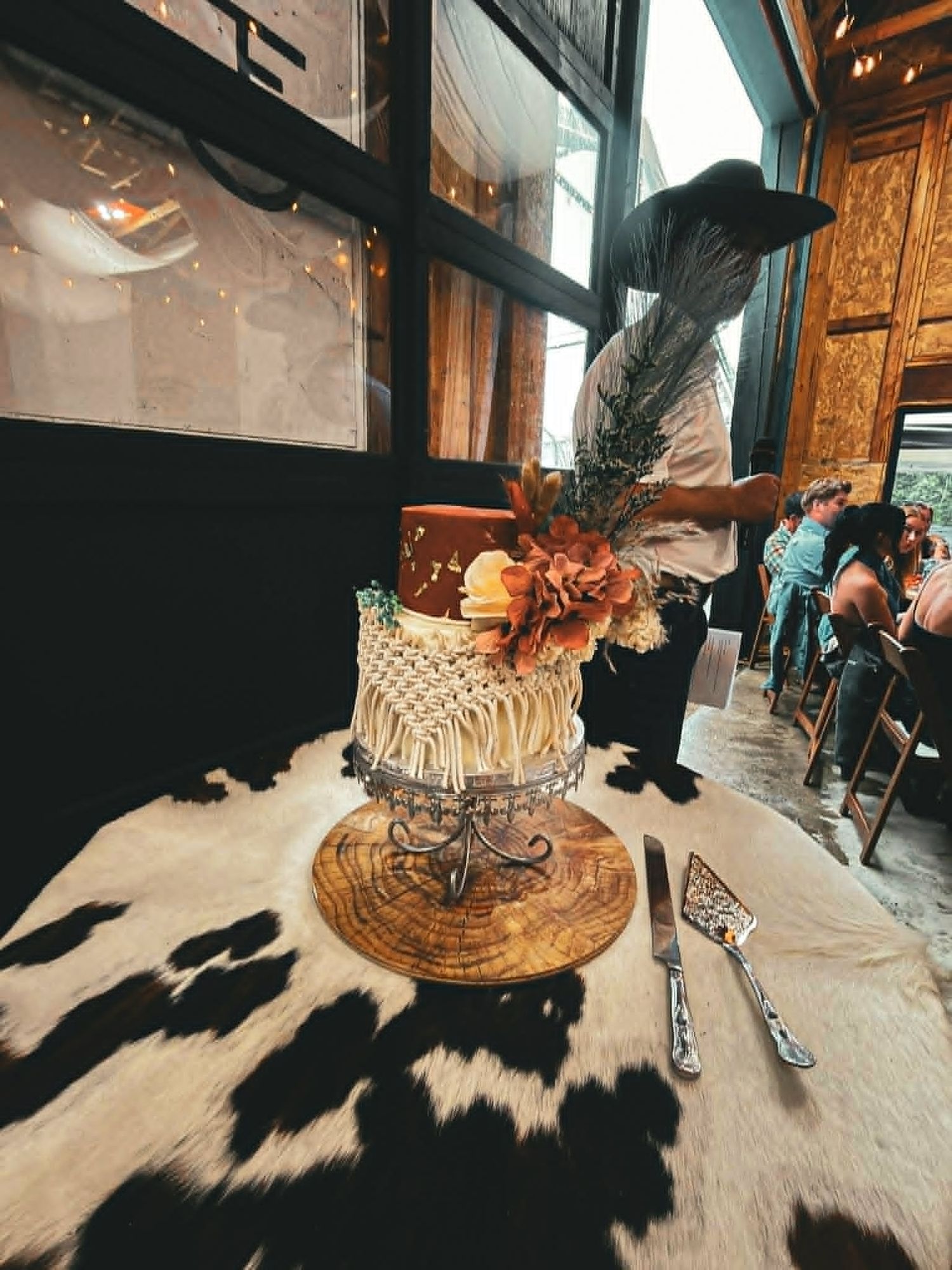 Rustic boho wedding cake with macrame and dried florals on wood slice stand, displayed on cowhide rug at western venue.