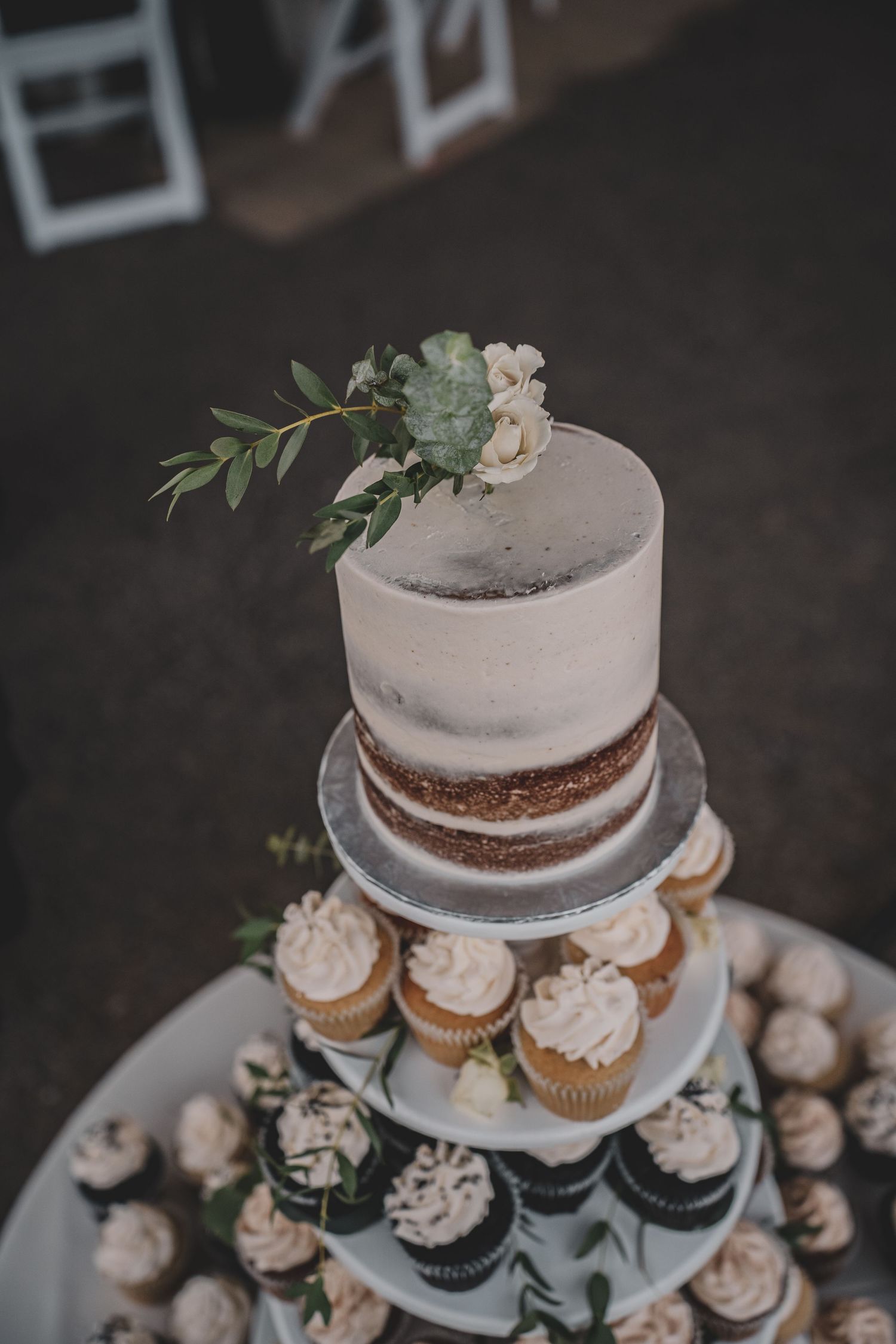 Elegant semi-naked wedding cake topped with greenery surrounded by cupcakes on tiered display stand at wedding, captured by  Free West.