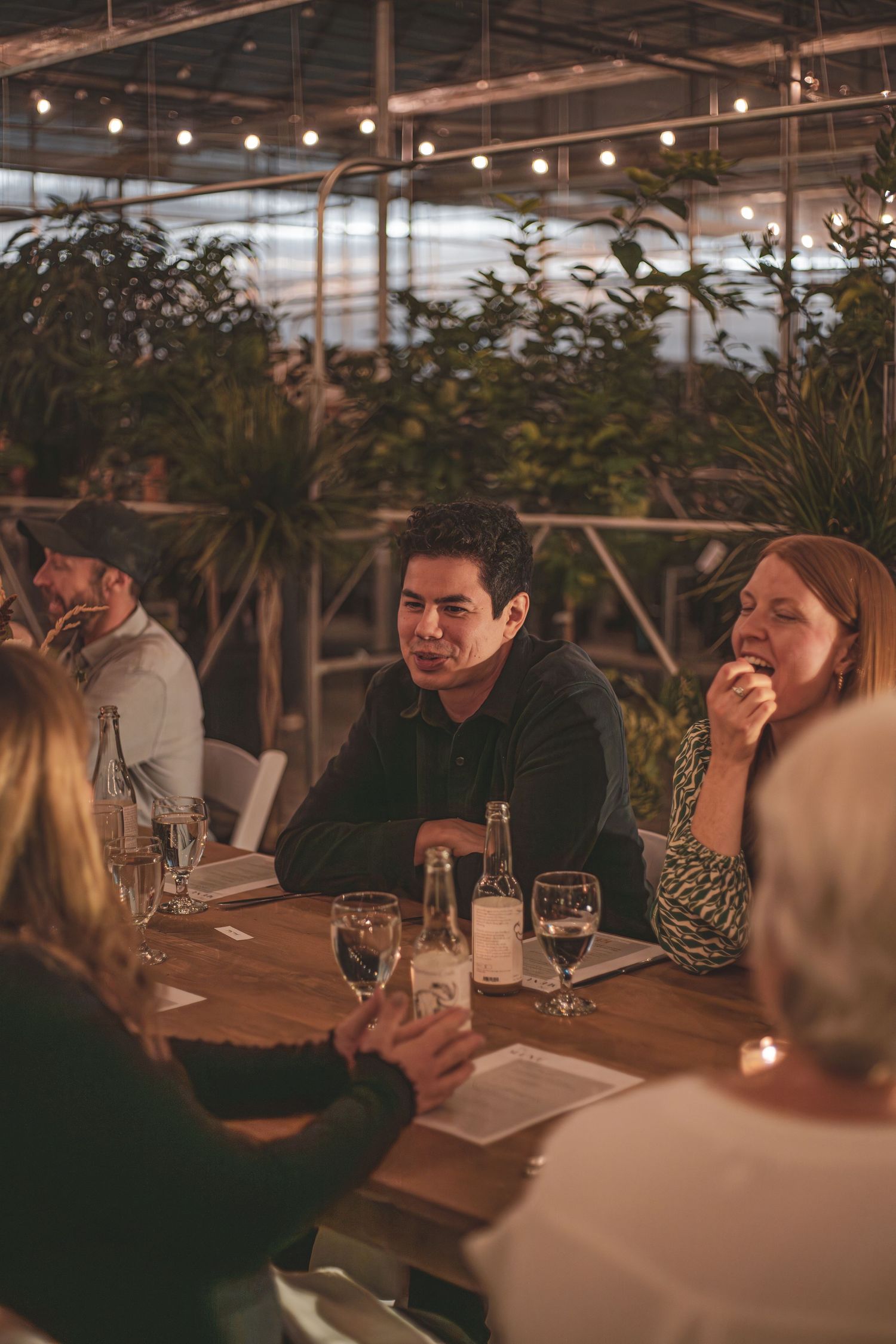 Guests seated at a rustic wooden table enjoy a dinner event inside a greenhouse with string lights above at a photography dinner event.