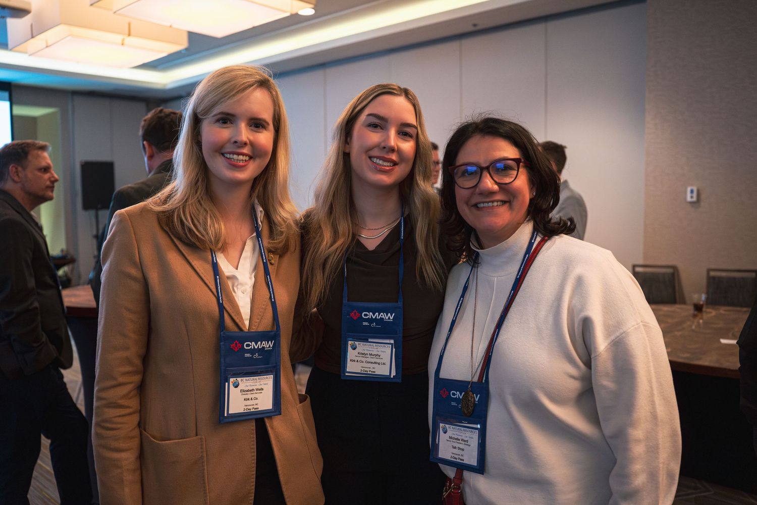 Three women wearing conference name badges smile together at a professional networking event.