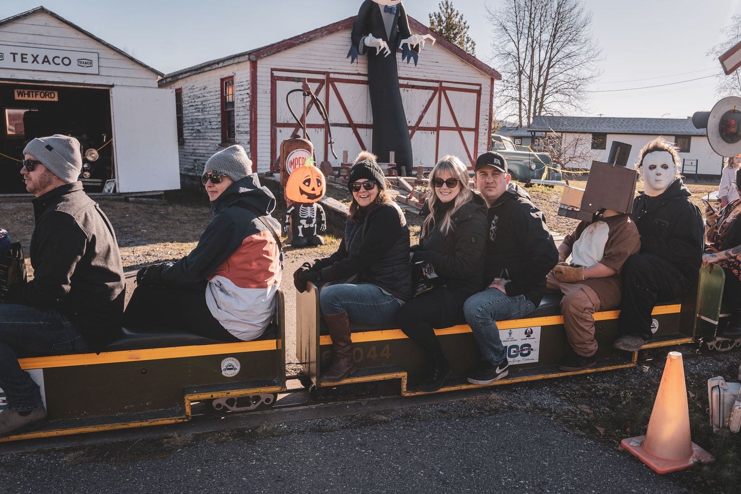 Group of people in Halloween costumes ride a flatbed train at an outdoor fall festival near a Texaco station.