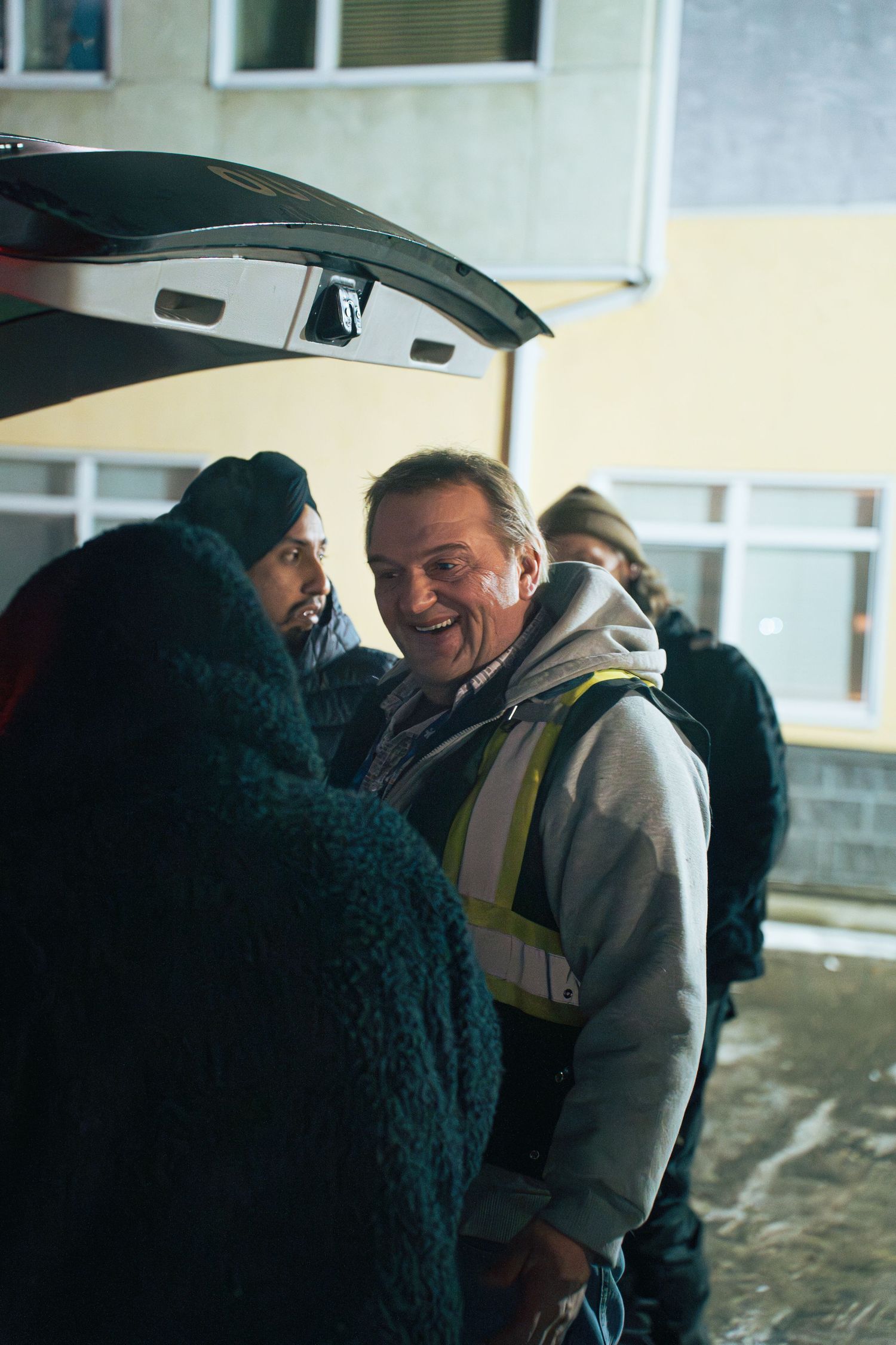 A smiling man in a safety vest stands near an open car trunk with others in a snowy outdoor setting at night.