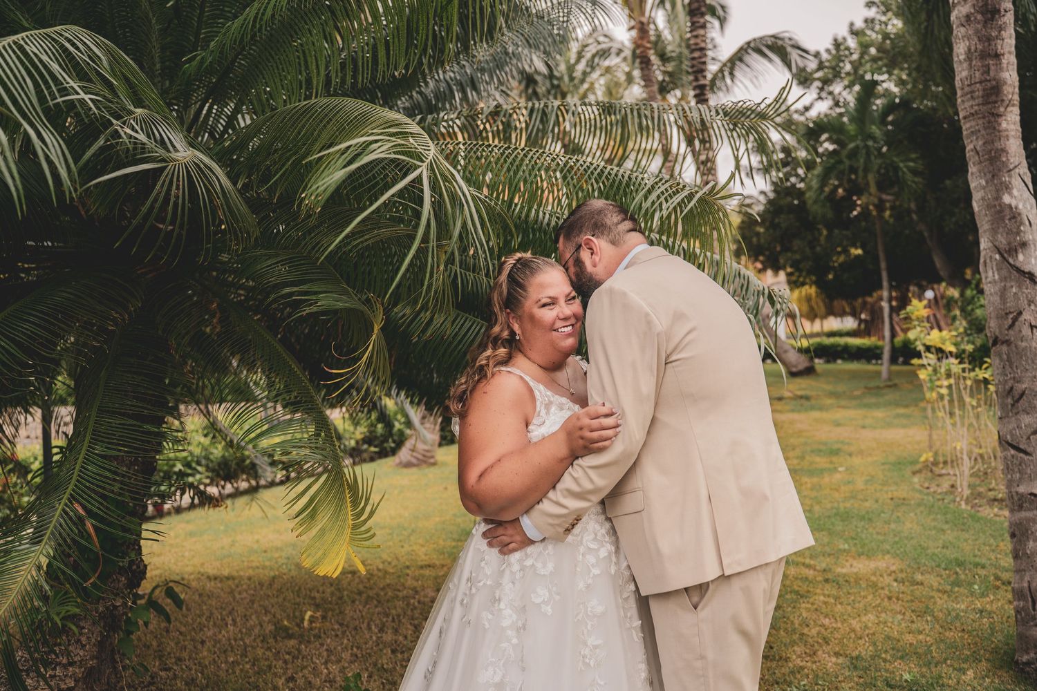 Happy bride in white lace gown embraces groom in tan suit beneath lush tropical palm trees outdoors.