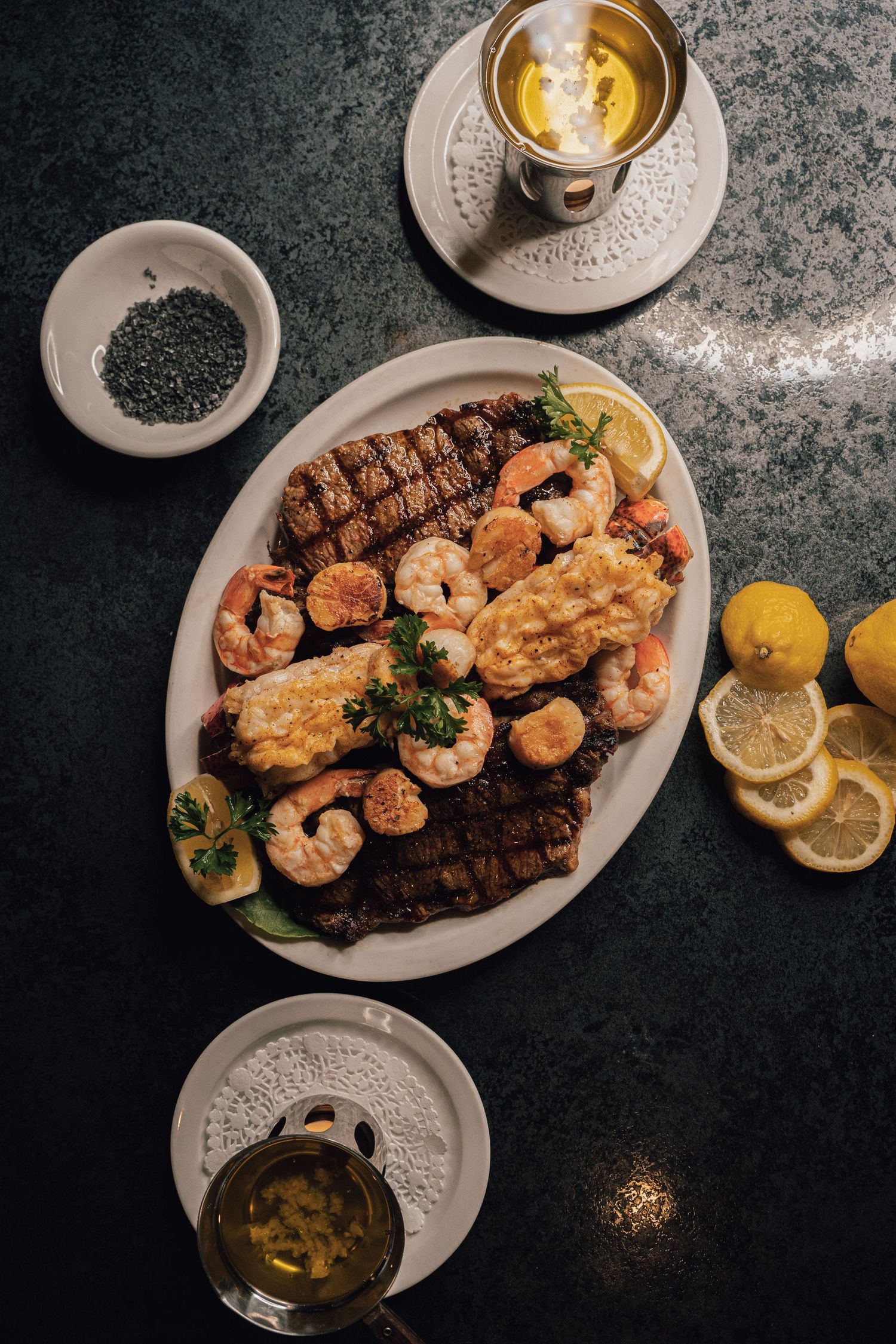 Elegant surf and turf platter with grilled steak, shrimp, lemon slices, and butter sauce on dark marble.