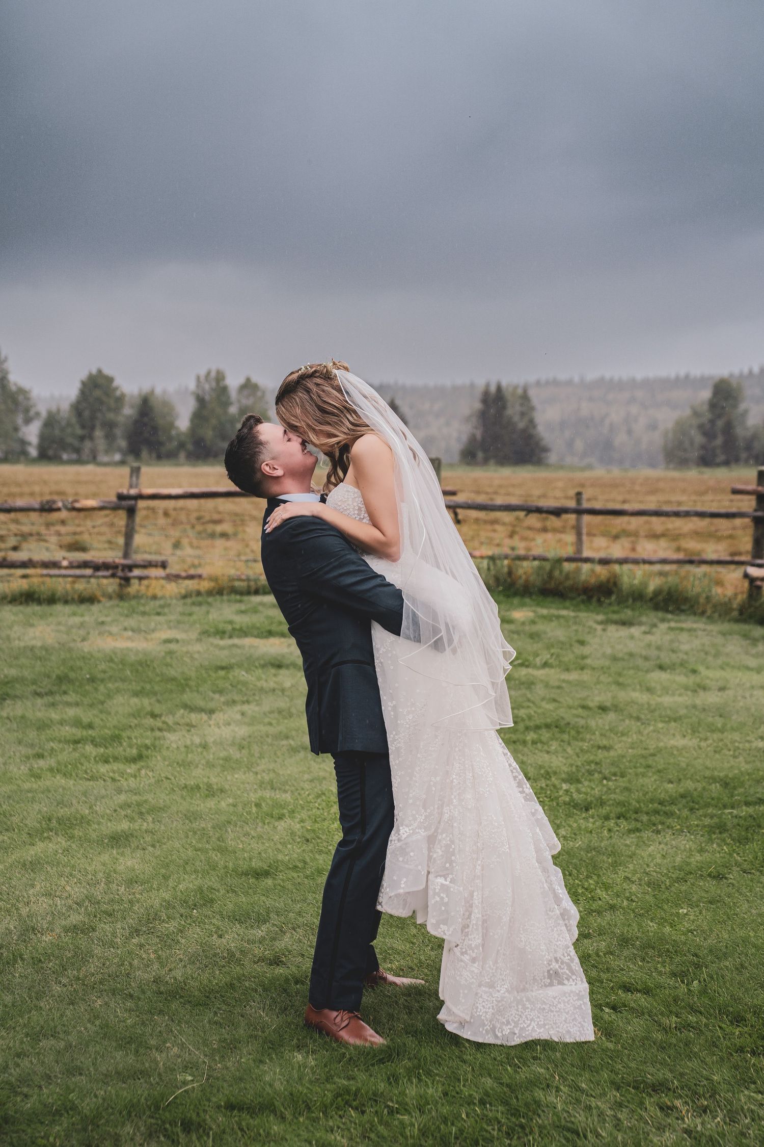 Groom lifting bride in white lace gown for a kiss on a green lawn under dramatic stormy skies.