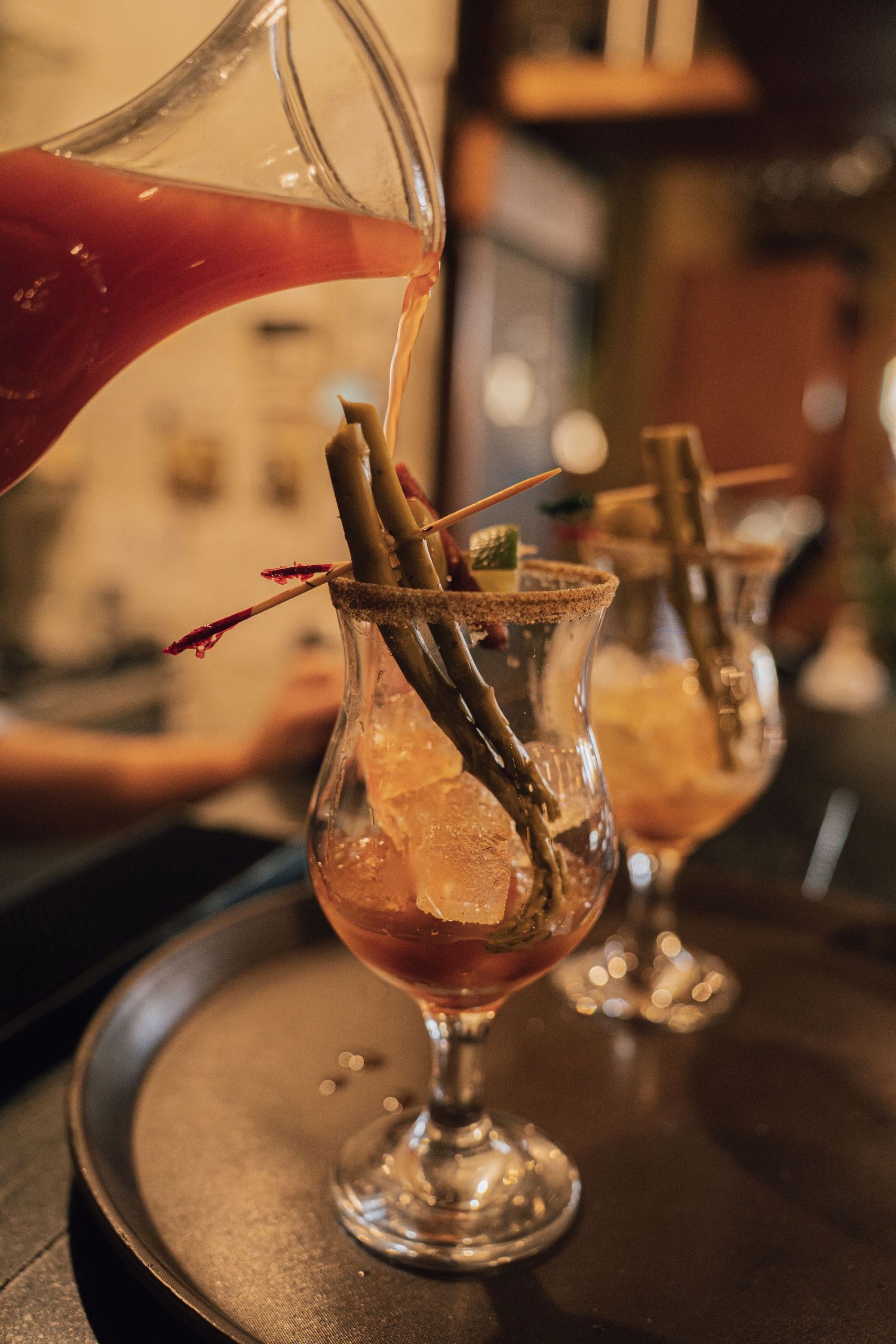 Red liquid being poured into ornate cocktail glasses garnished with cinnamon sticks on a tray.