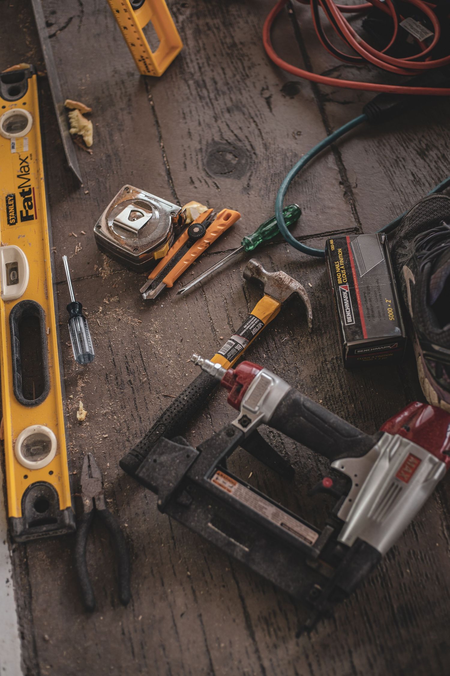 Carpentry tools including nail gun, level, hammer, and tape measure laid out on wooden floor.