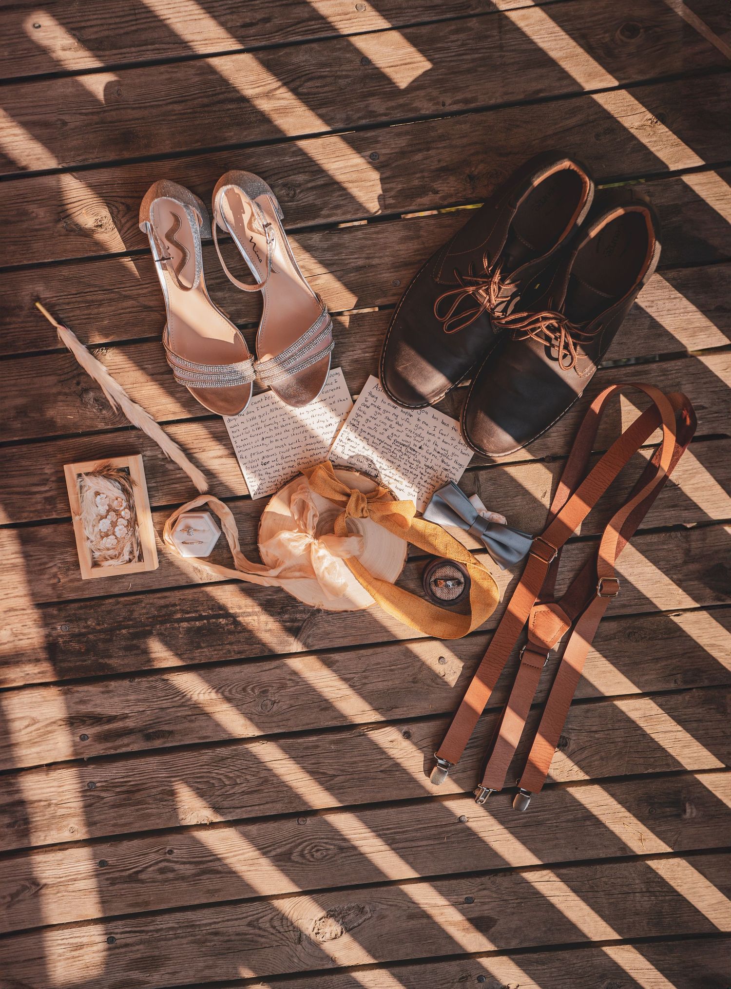 Flat lay of wedding accessories including bridal heels, groom boots, suspenders, and stationery on wood.