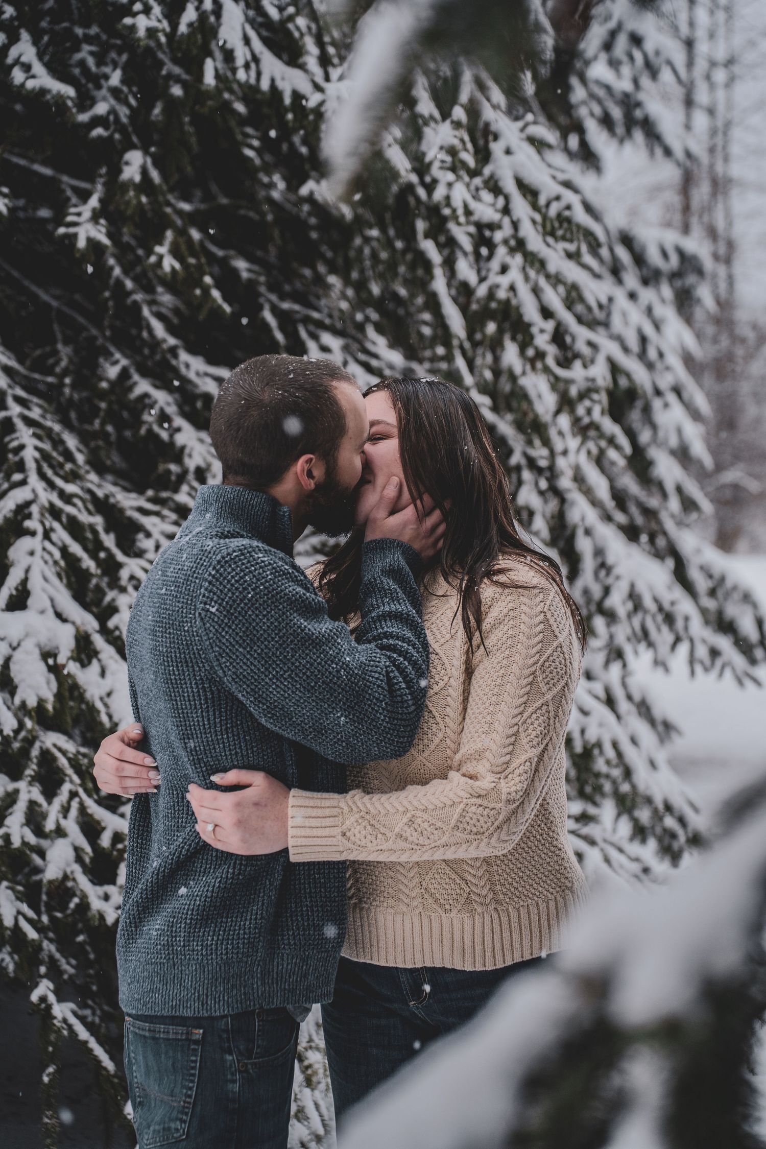 Romantic couple kissing and embracing in snowy winter forest surrounded by snow-covered pine trees.