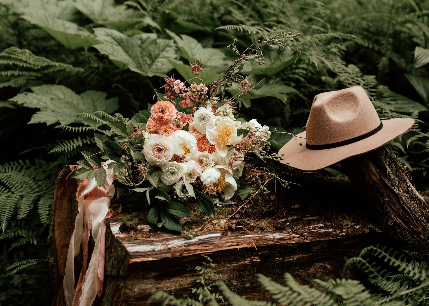 Bridal bouquet of white and peach flowers resting beside a tan hat among lush green ferns details captured at elopement  at ancient forest.