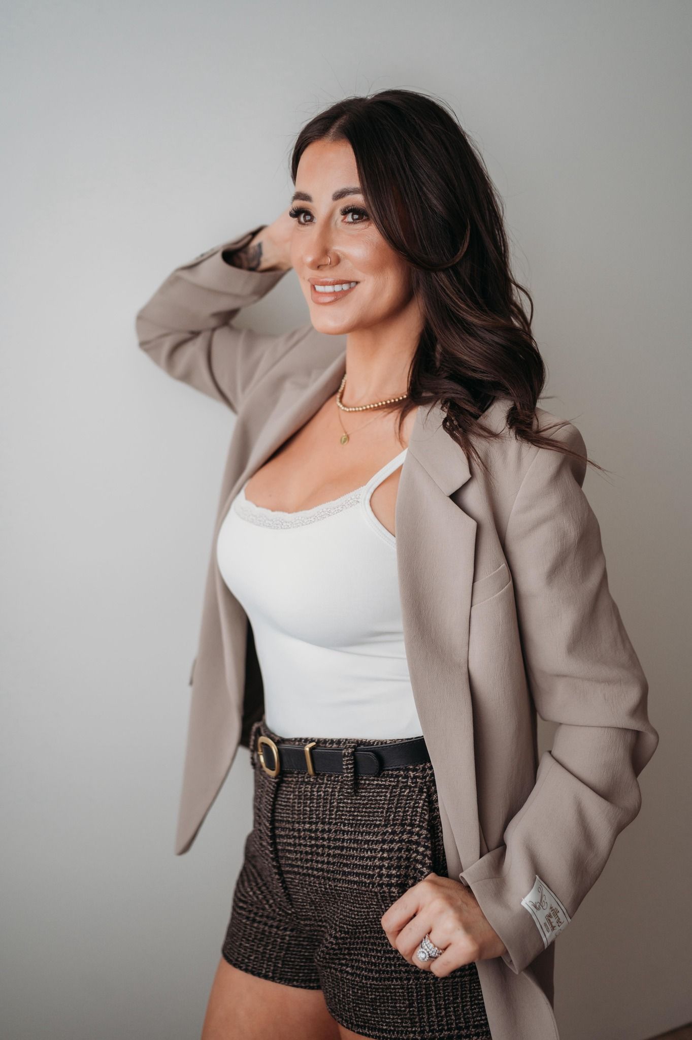 Confident brunette woman smiling in white tank top, blazer, and plaid shorts against neutral wall.